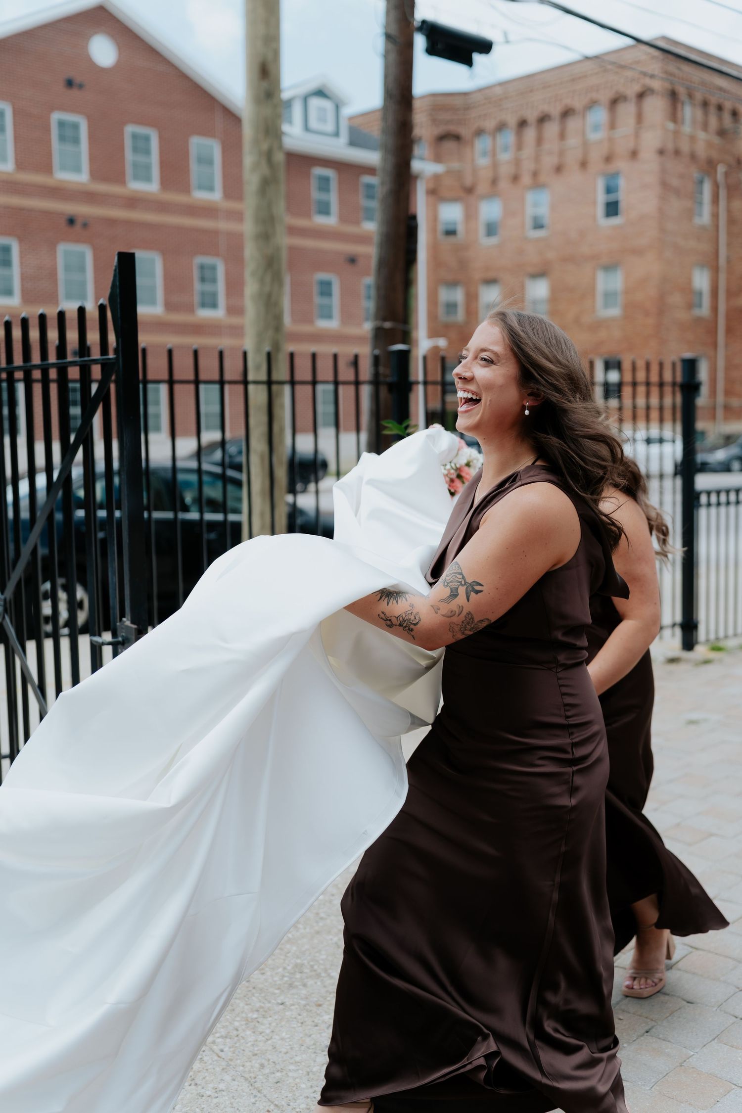 Bridesmaid in dark brown gown laughing while holding white wedding dress train on urban sidewalk.