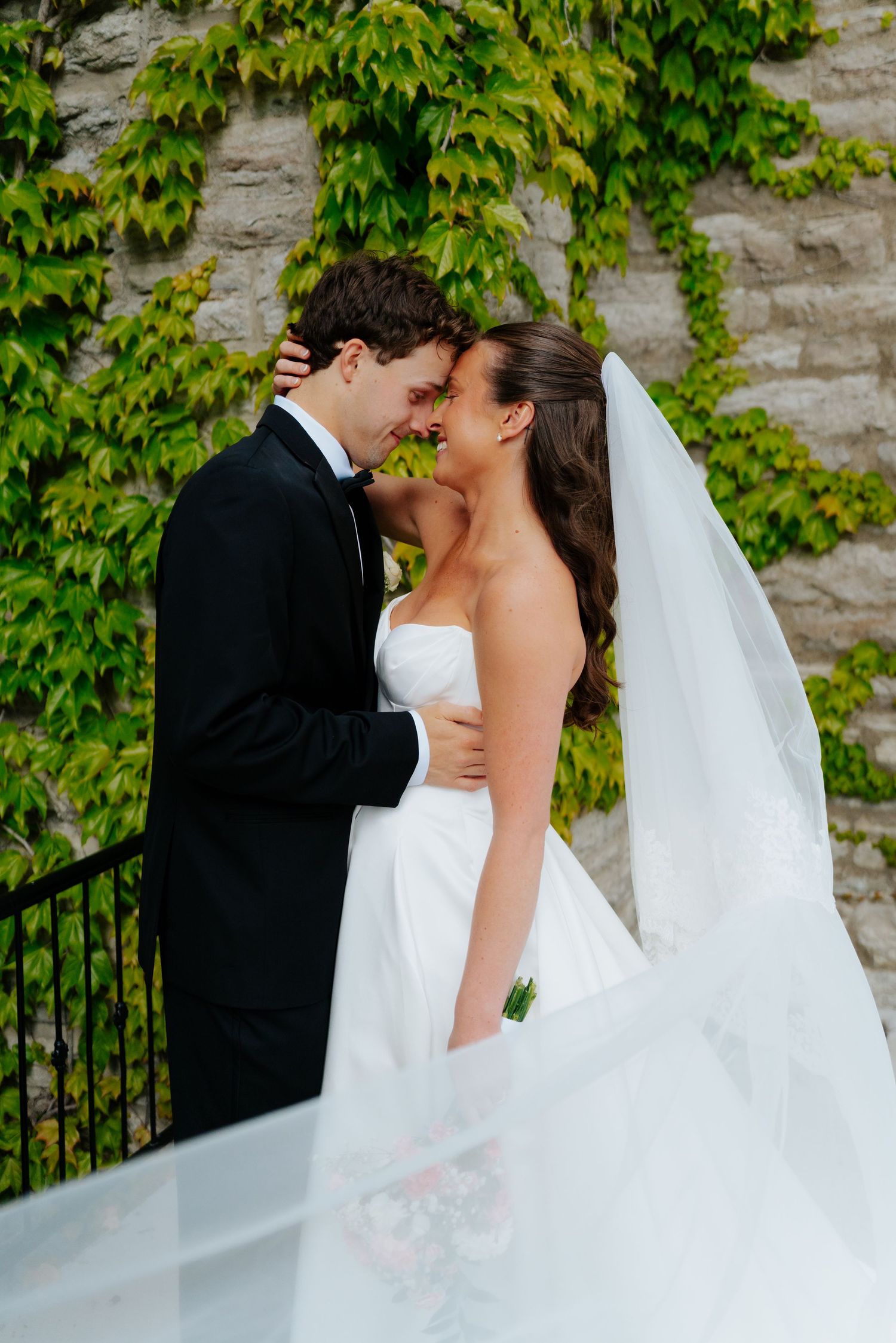 Bride and groom share a romantic moment in front of an ivy-covered stone wall, with her white gown and veil flowing.