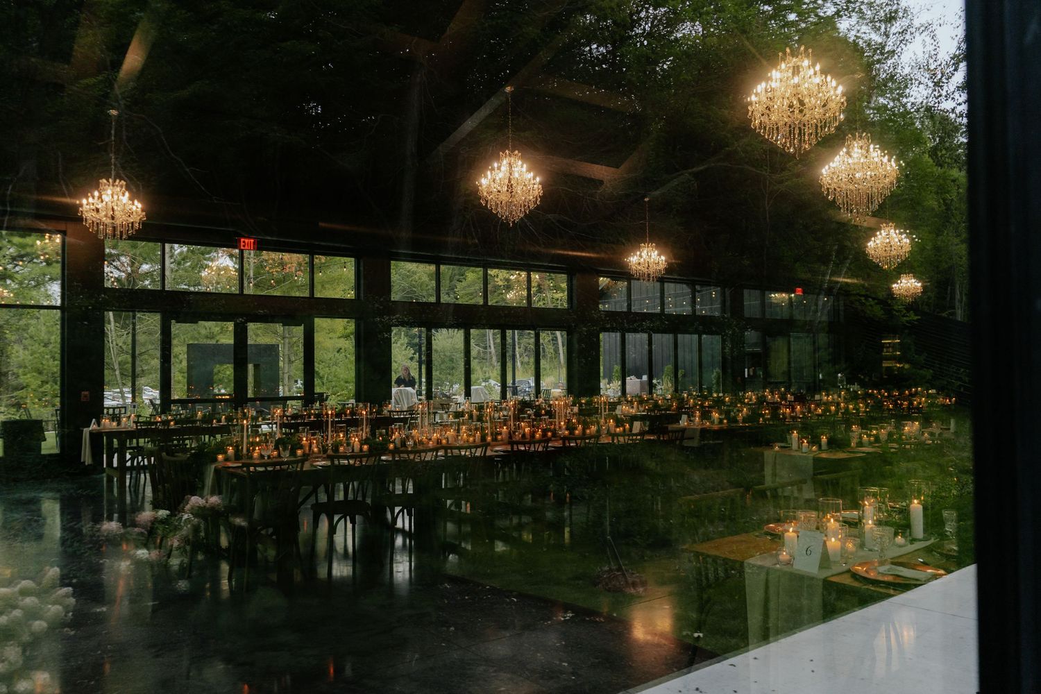 Elegant wedding reception hall with long banquet tables, glowing chandeliers, and lush garden views through floor-to-ceiling windows.