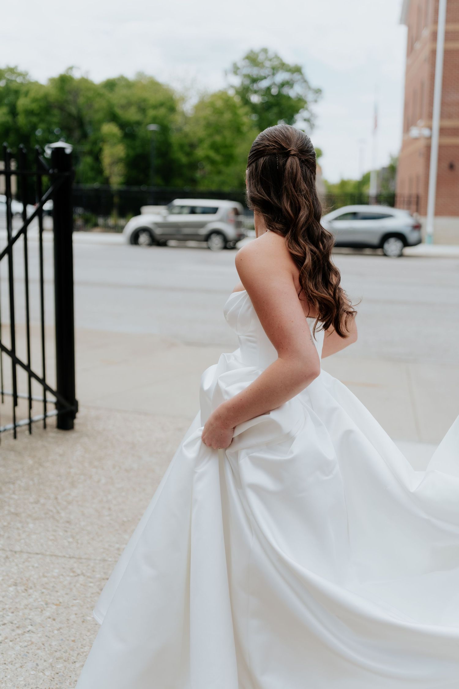 Bride in strapless white ballgown holding her dress while standing near a black iron gate on a city street.