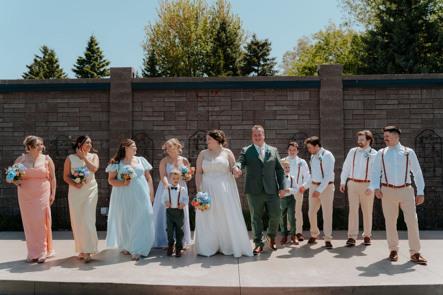 Wedding party poses outdoors in front of brick wall, bridesmaids in blue and pink, groomsmen in green suits with suspenders.