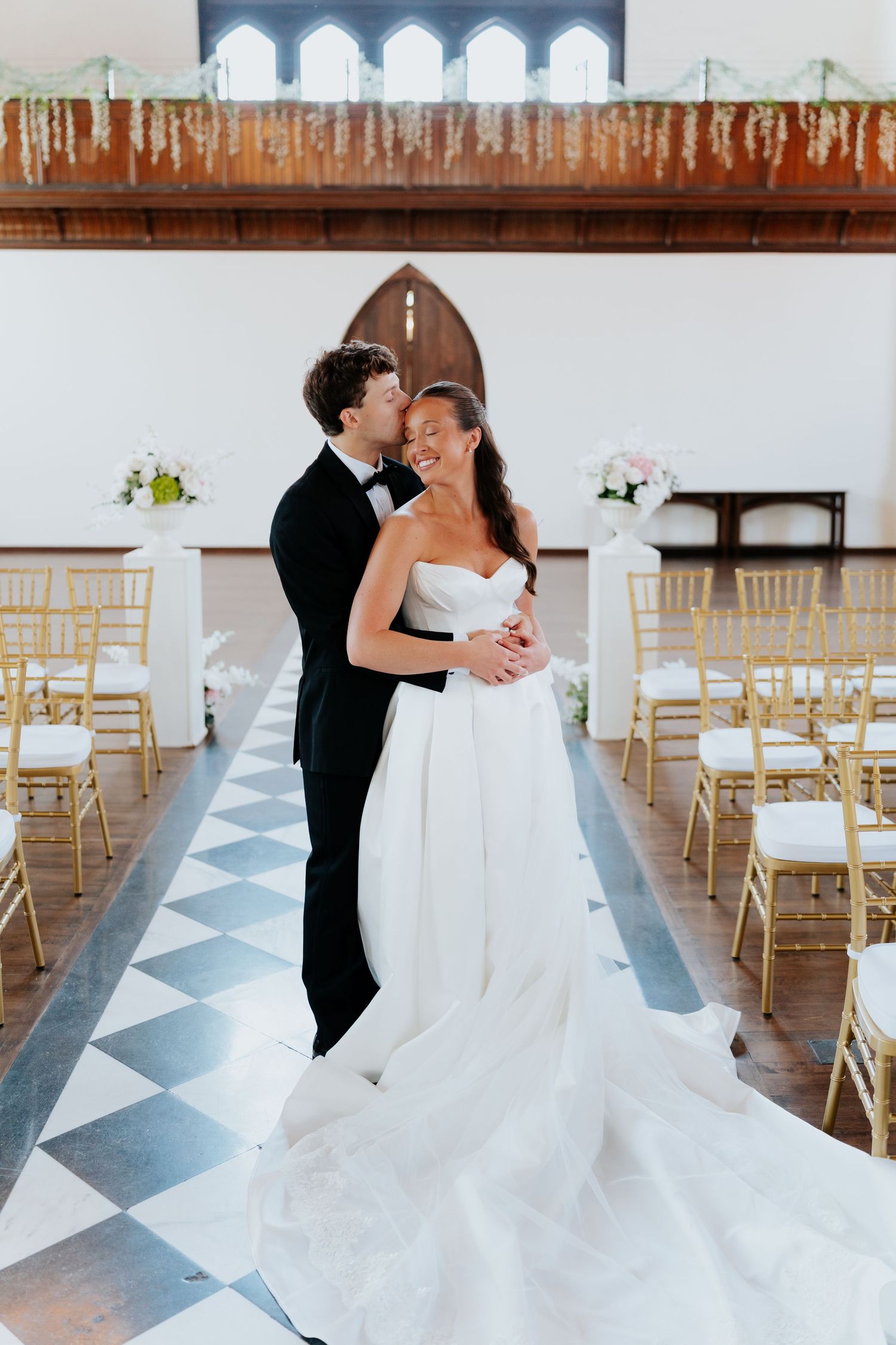 Bride and groom embrace in elegant church aisle with gold chairs, checkered floor, and floral decorations on their wedding day.