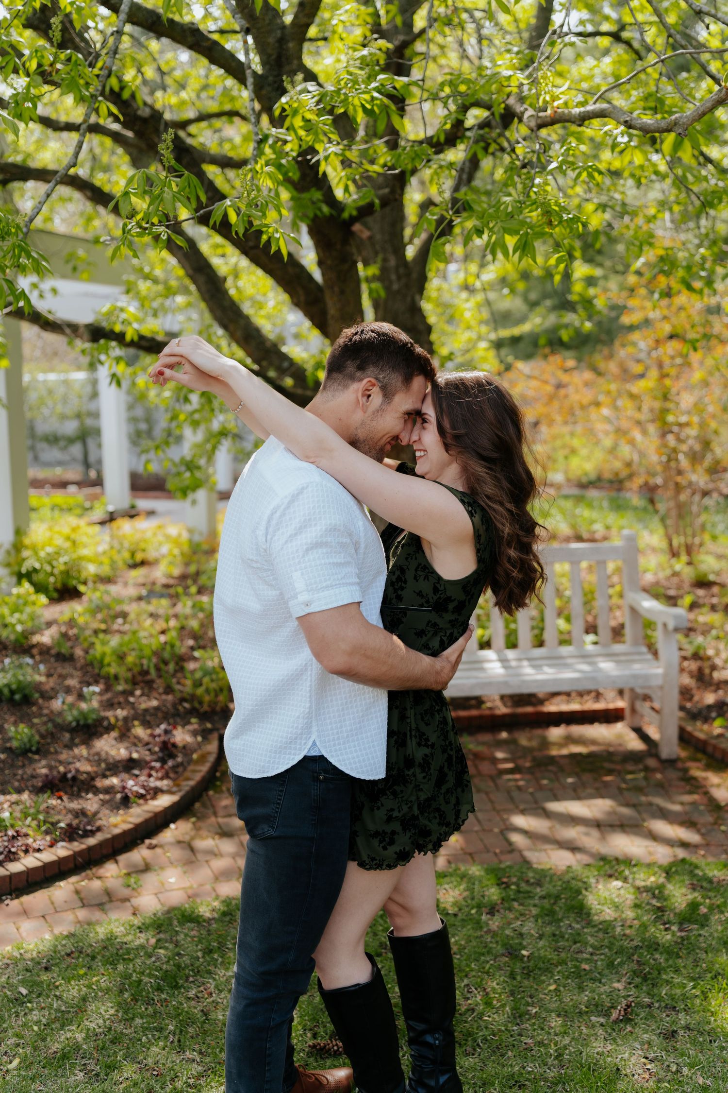 Romantic couple dancing and embracing under a tree in a lush garden with a white bench backdrop.