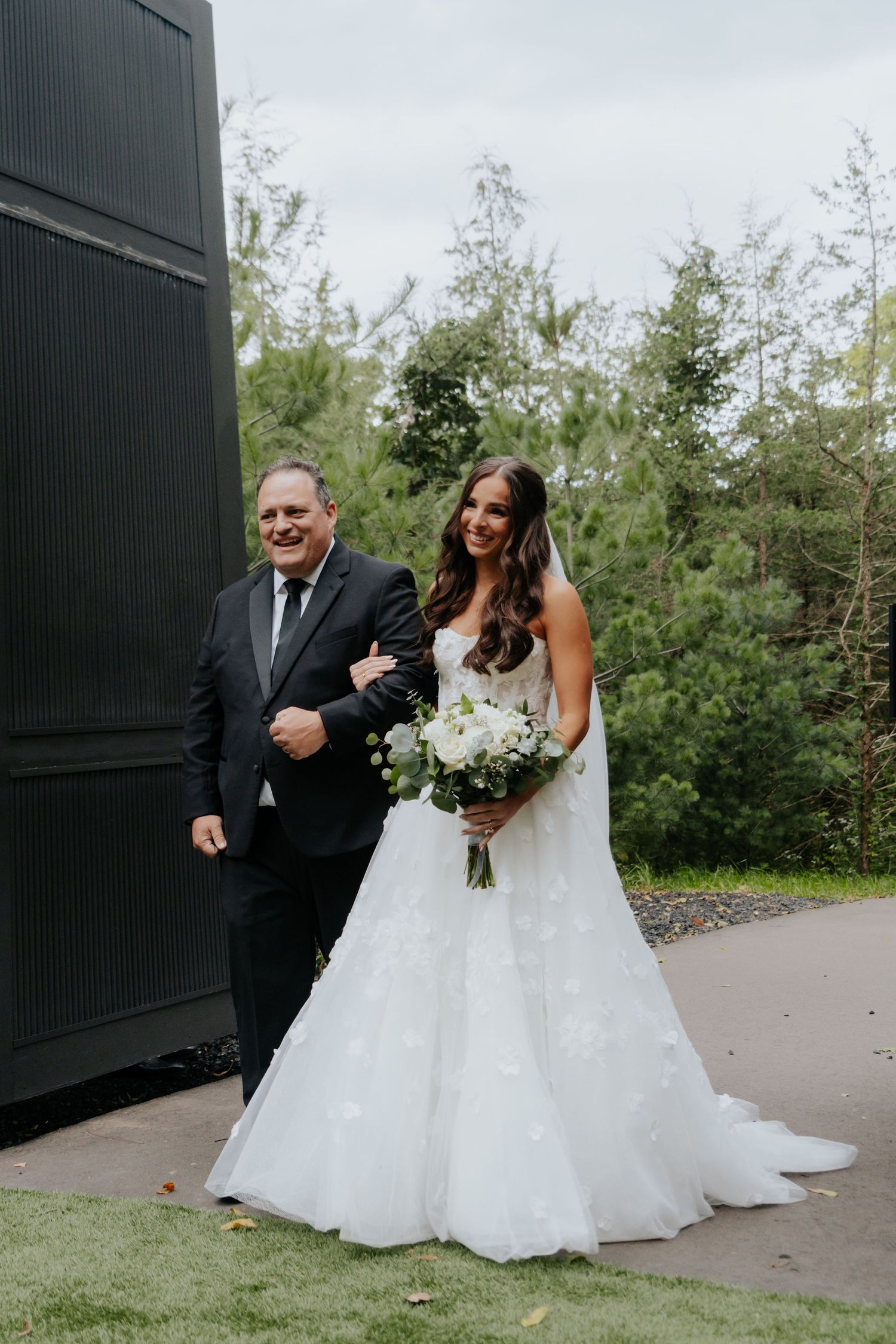 Bride in white floral gown holding bouquet walks with suited man outdoors at wedding venue surrounded by greenery.