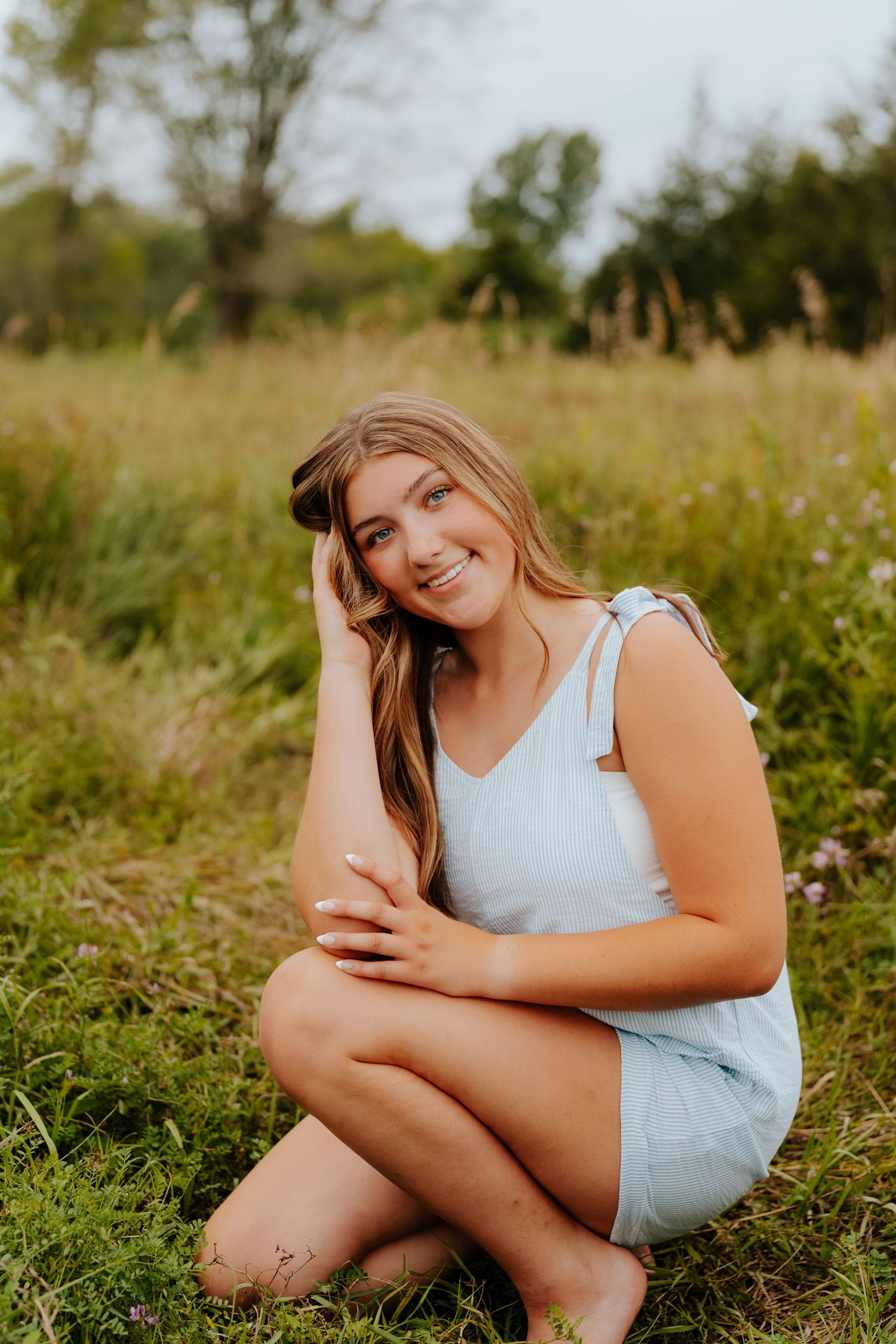 Young woman in light blue romper sitting in a grassy field, smiling warmly during a golden hour outdoor photo session.