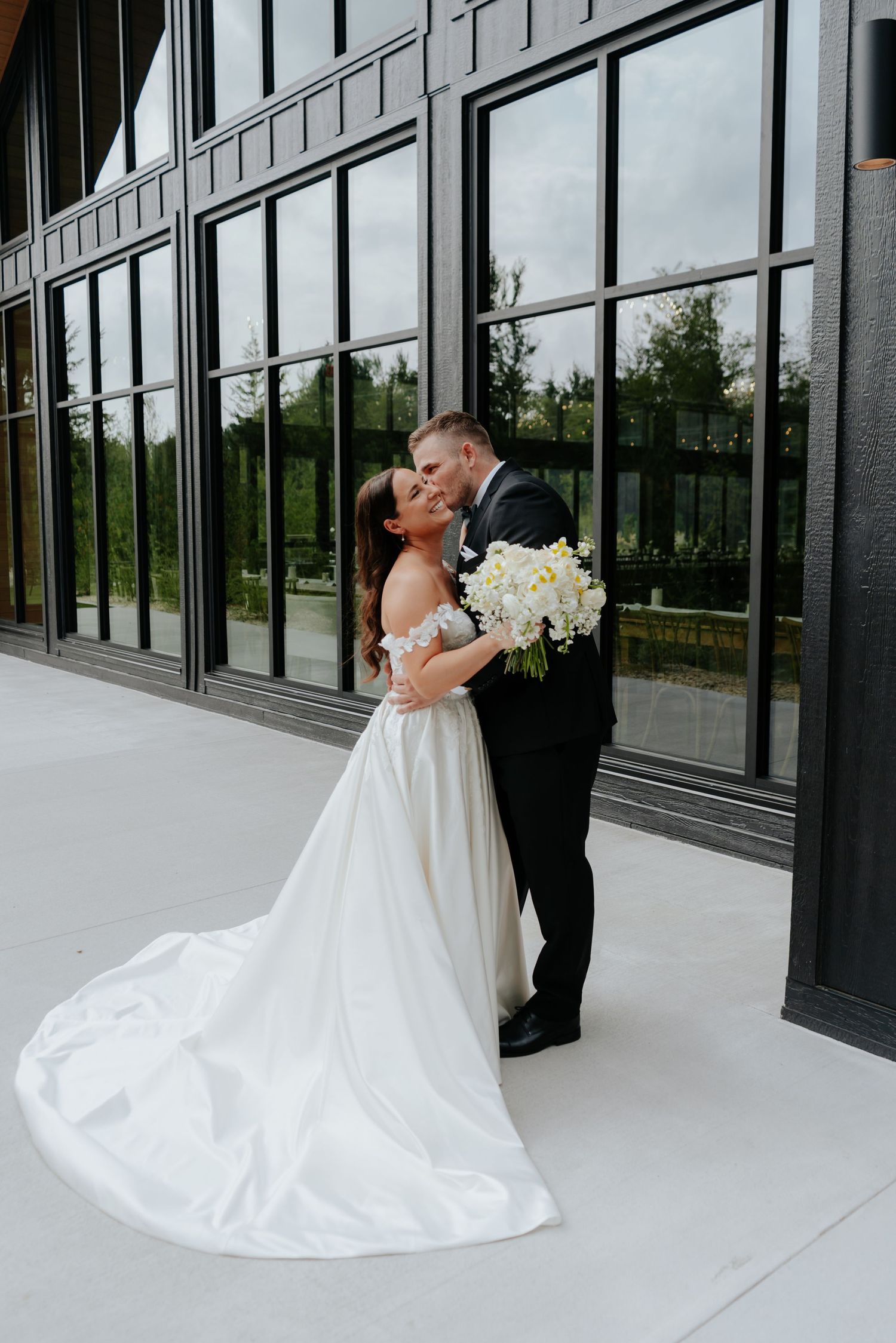 A bride and groom share a romantic kiss outdoors, with the bride holding a white bouquet and wearing an elegant off-shoulder gown.