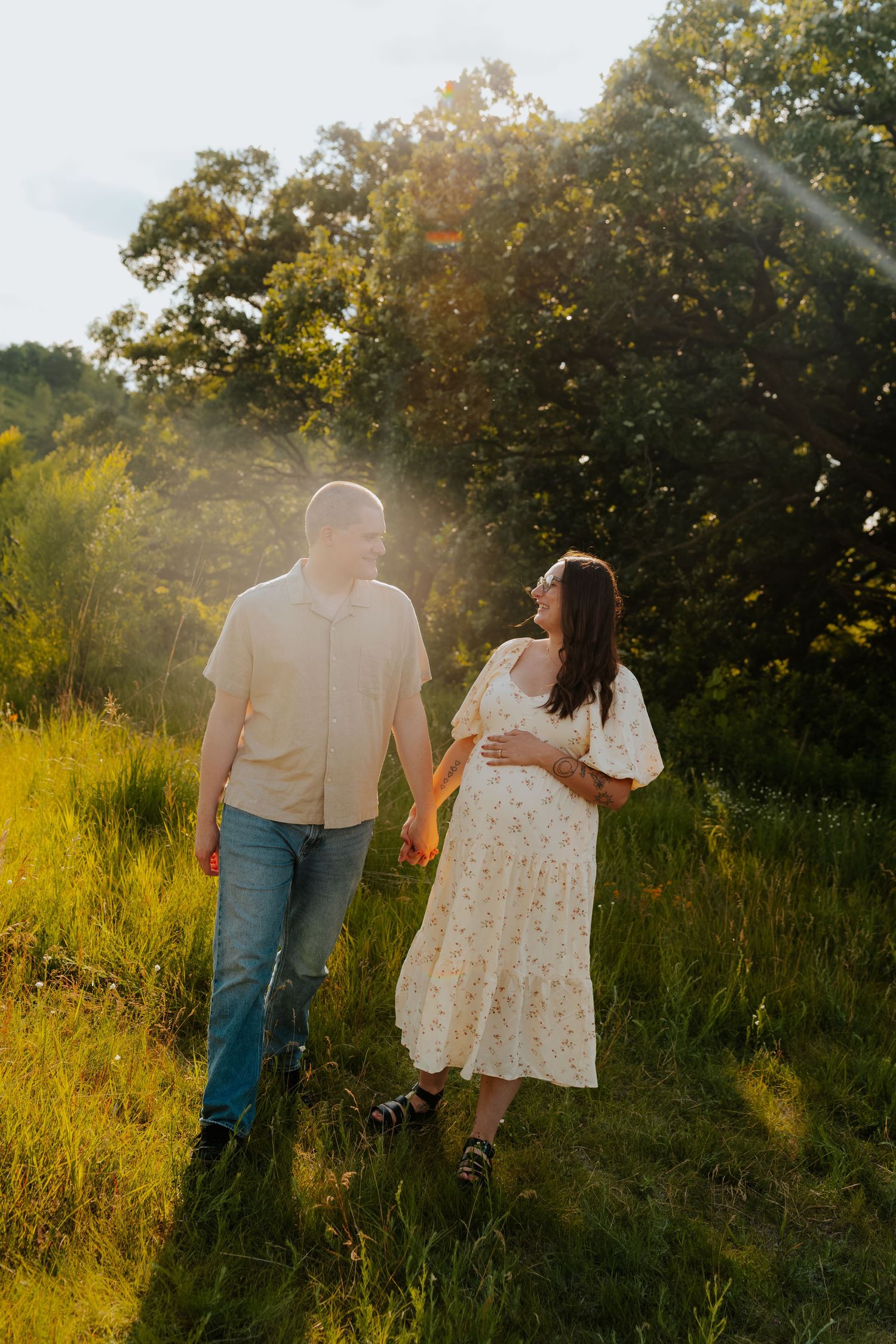 A couple holds hands walking through a sunlit green meadow, the woman in a floral dress cradling her baby bump.