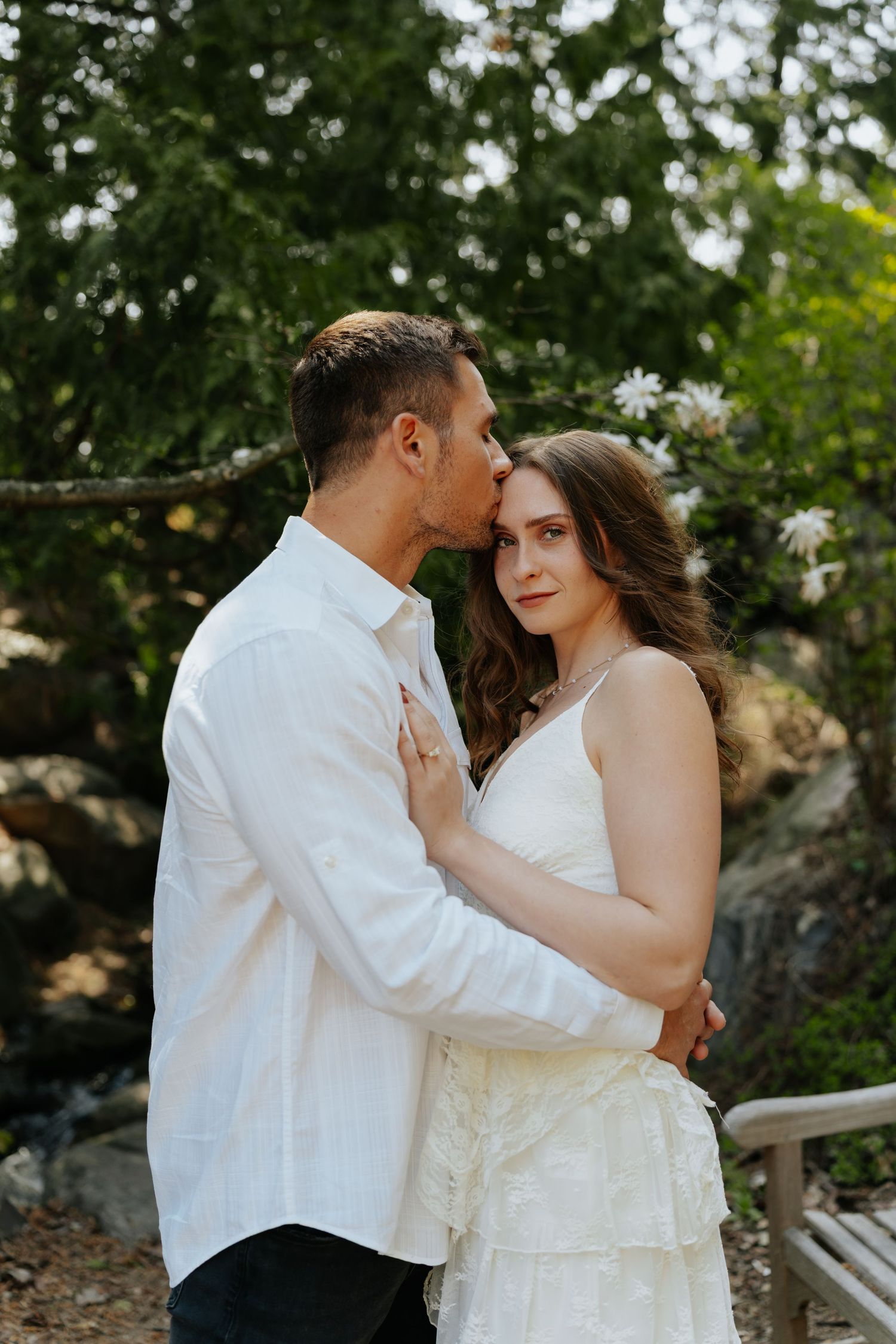 A couple in white attire shares a tender kiss outdoors, surrounded by lush greenery and blooming flowers.