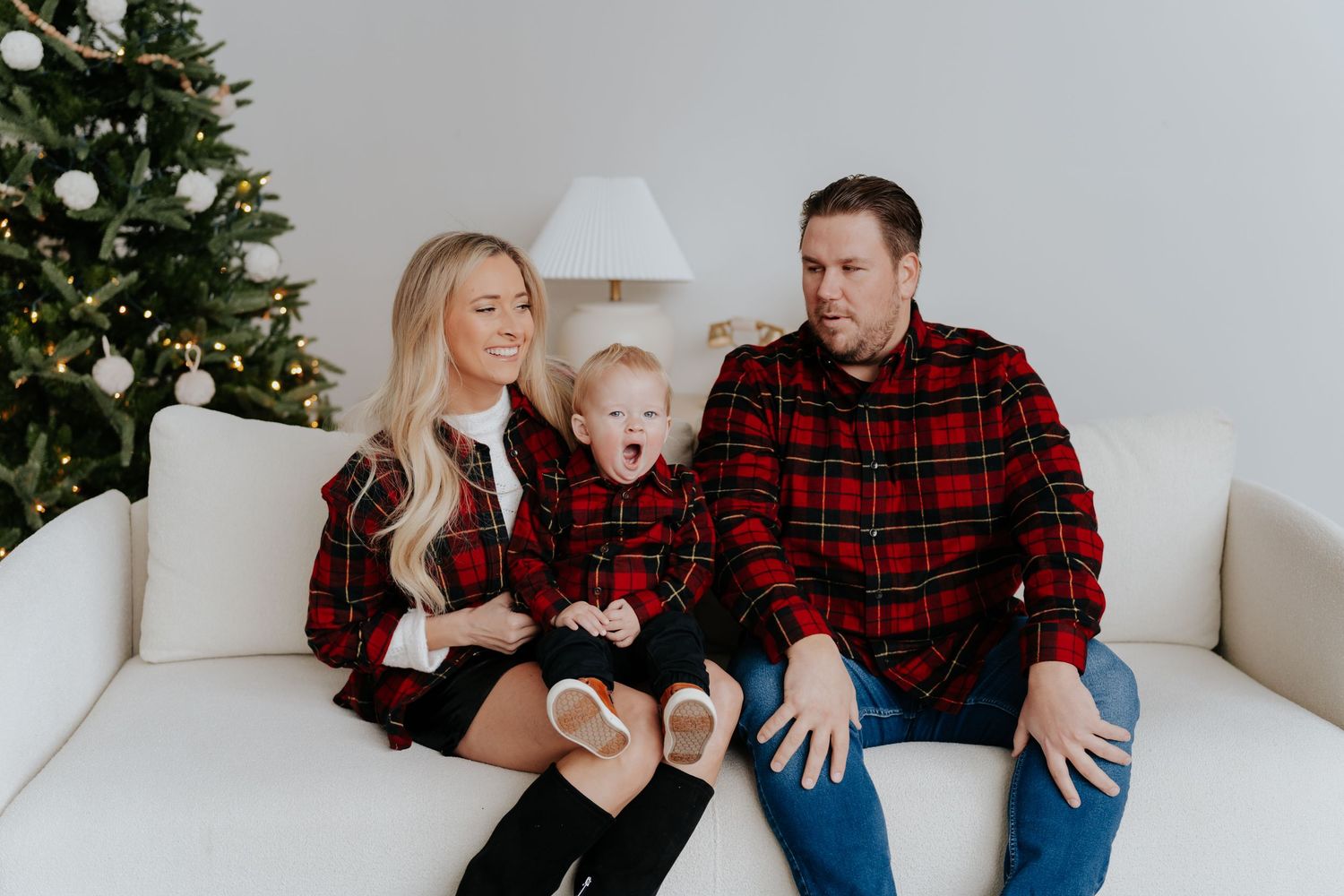 Family of three in matching red plaid shirts sitting on white sofa near Christmas tree for holiday photo.