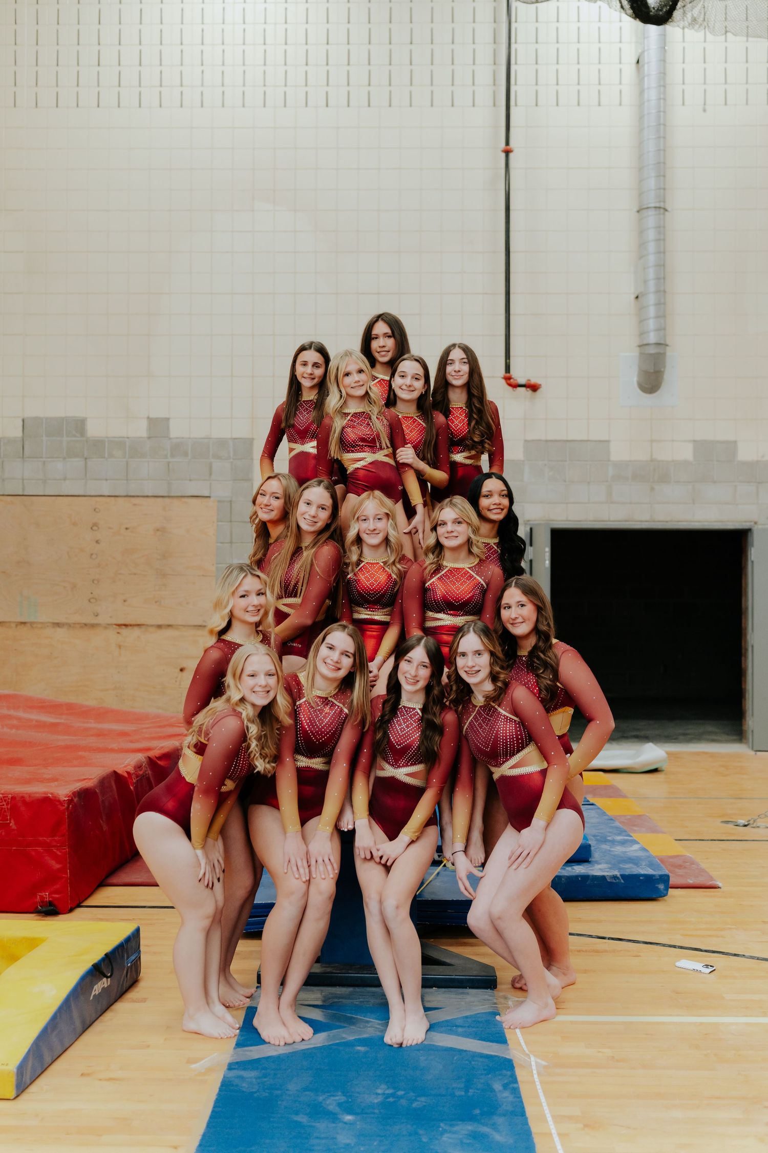 A cheerleading squad in red uniforms poses in a pyramid formation on gym mats indoors.