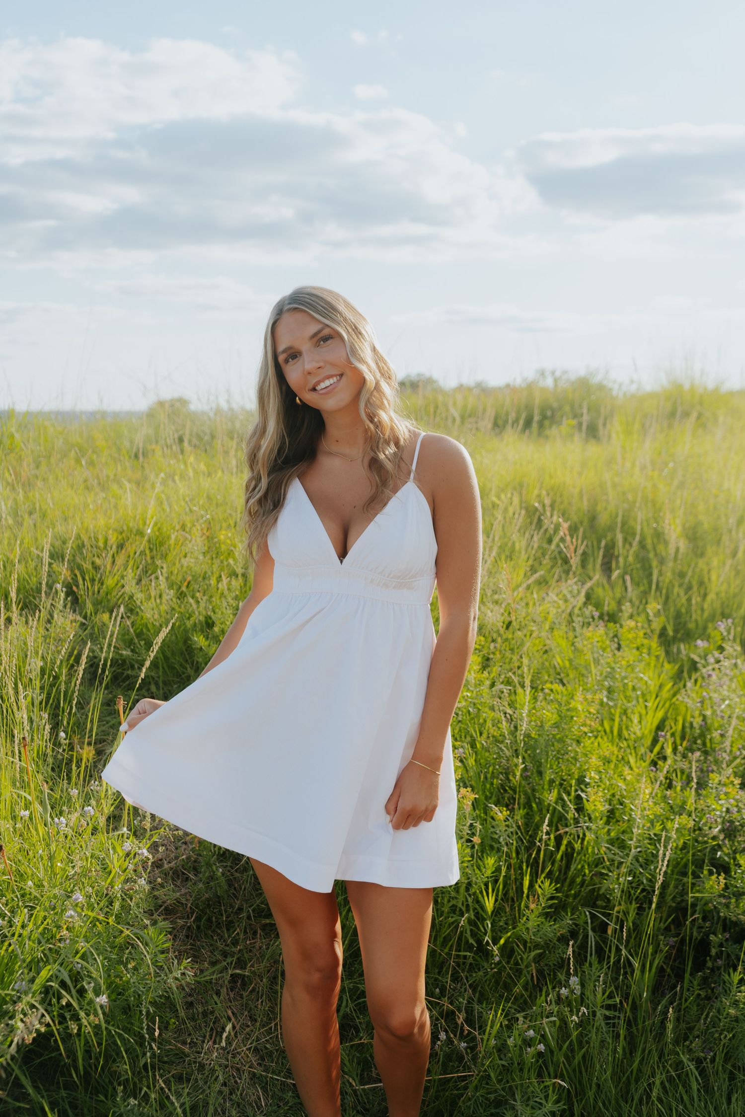 Smiling blonde woman in white sundress standing in lush green field under blue sky.