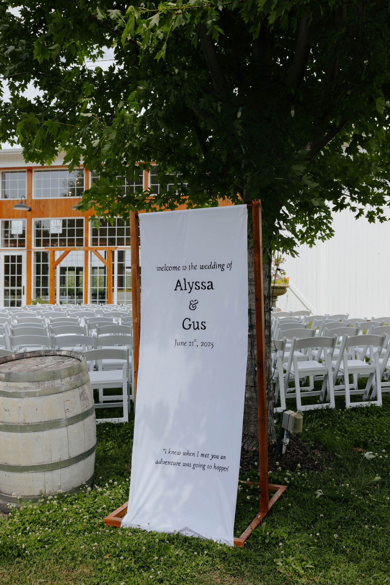 Outdoor wedding welcome sign for Alyssa and Gus, June 2023, displayed on wooden stand beside barrel with white chairs.