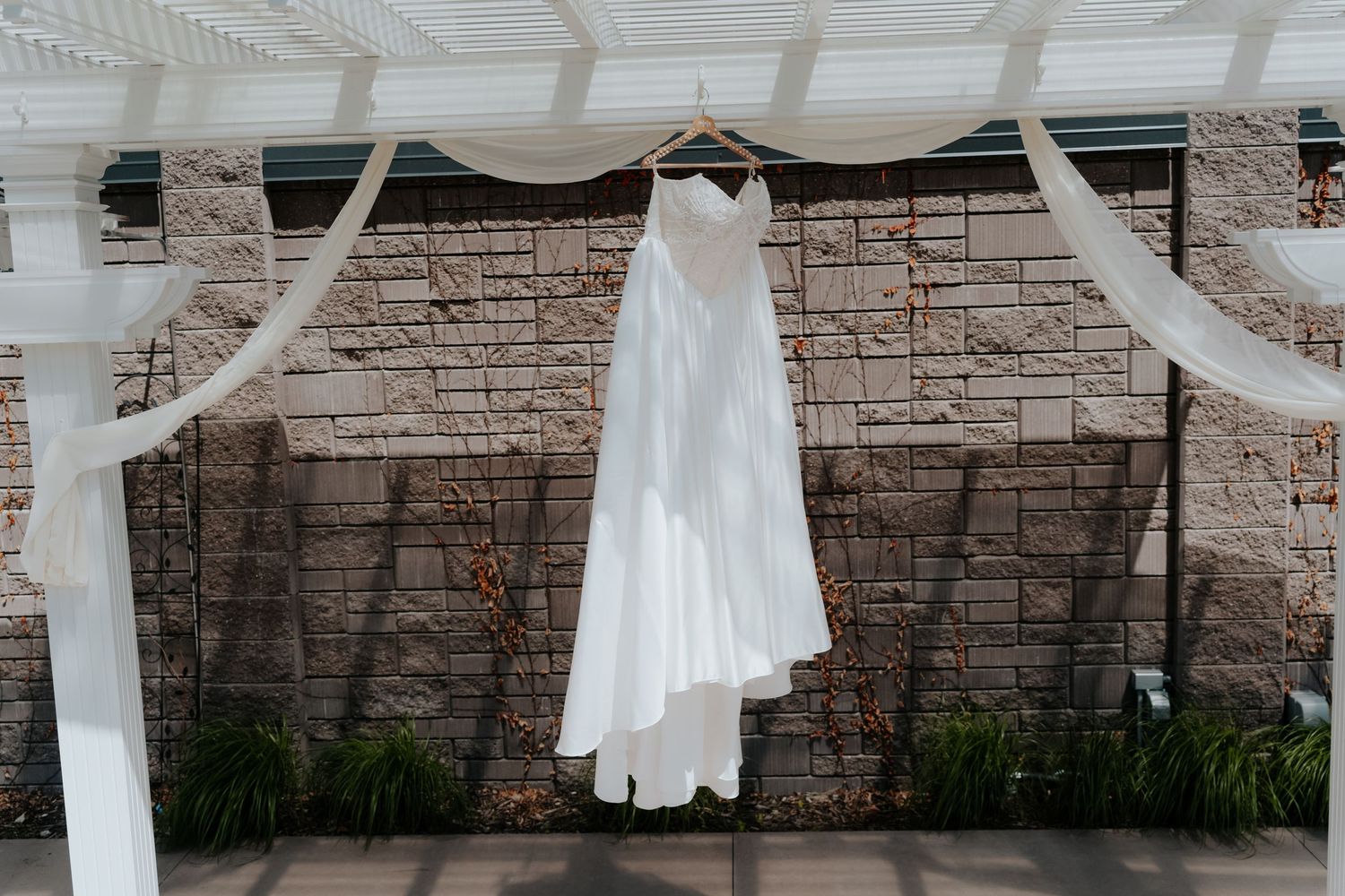 White wedding dress hanging on a hanger under a pergola with sheer draping against a brick wall.