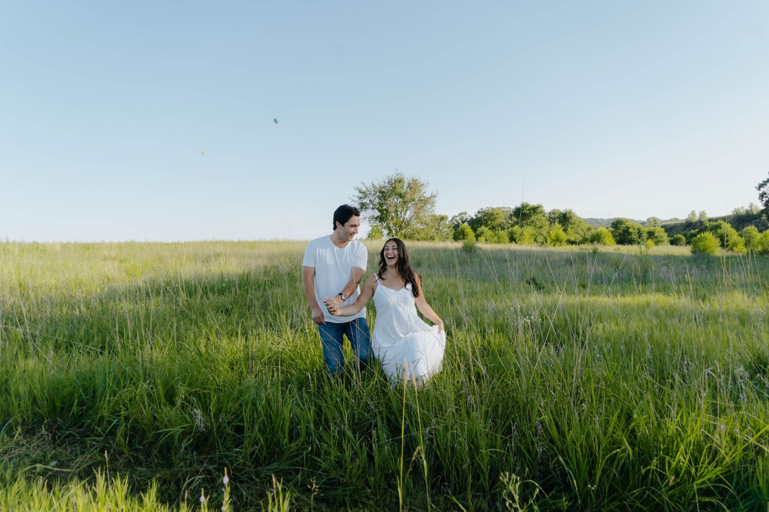 A couple stands together in a scenic green meadow during golden hour with sunlight illuminating the tall grass.