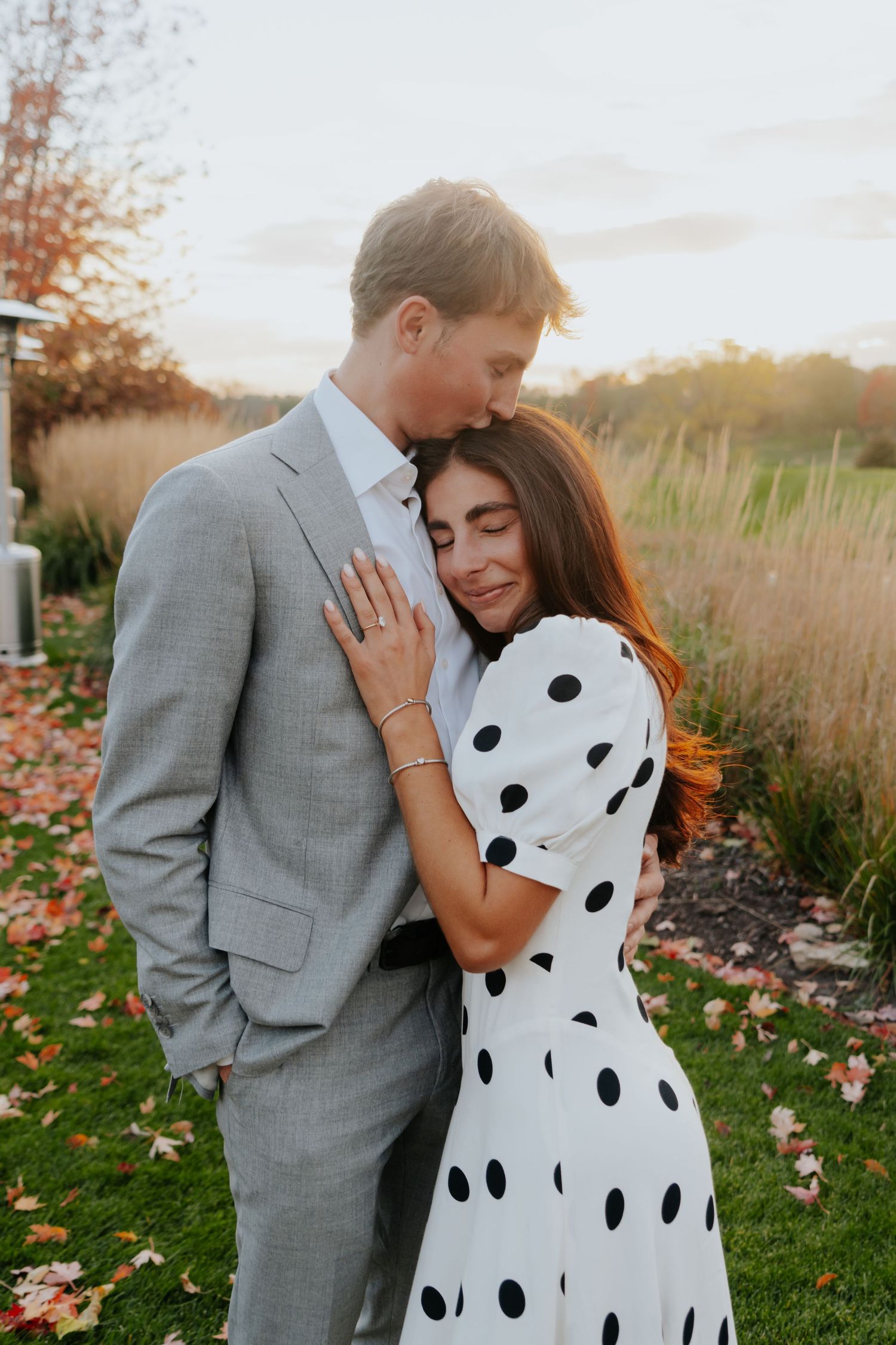 Couple in grey suit and polka dot dress share an intimate moment among autumn leaves.