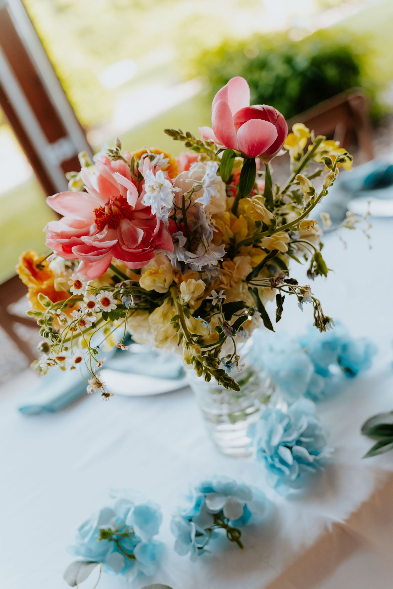 Elegant wedding table centerpiece featuring pink tulips, yellow blooms, and blue paper flowers on white linen.