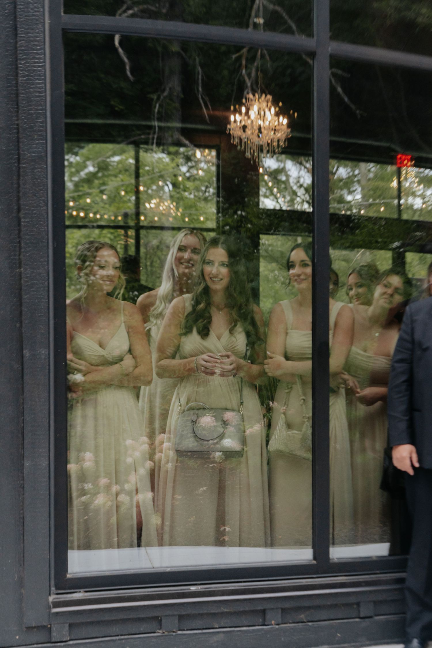 Bridesmaids in sage green dresses gather behind a large window with a chandelier, creating an elegant reflection effect.