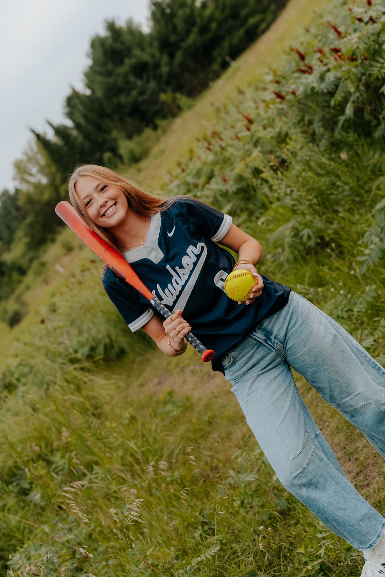 Smiling softball player in Hudson jersey holding red bat and yellow ball in a lush green field.