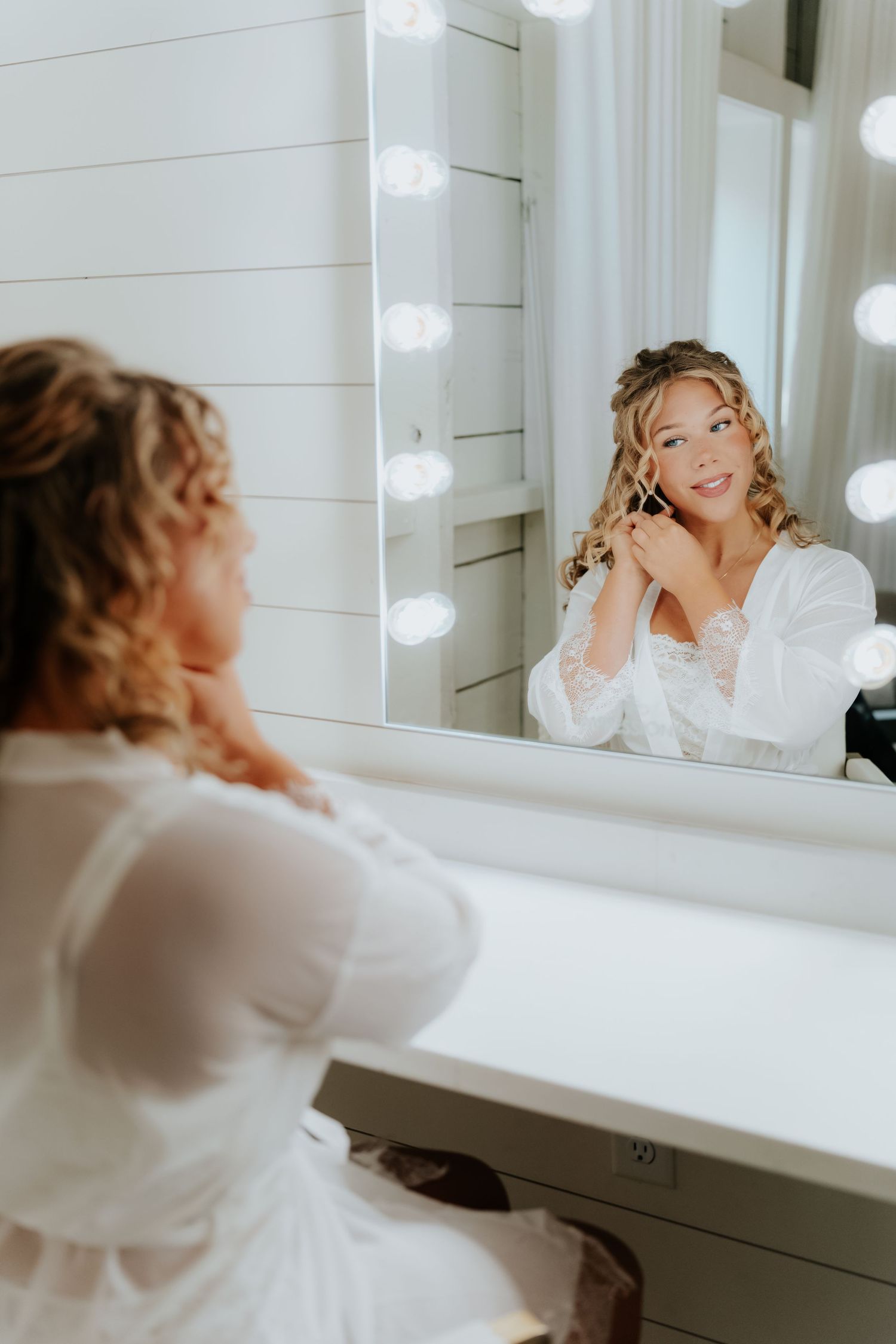 Bride in white robe applying earrings at a Hollywood mirror vanity while getting ready for her wedding day.