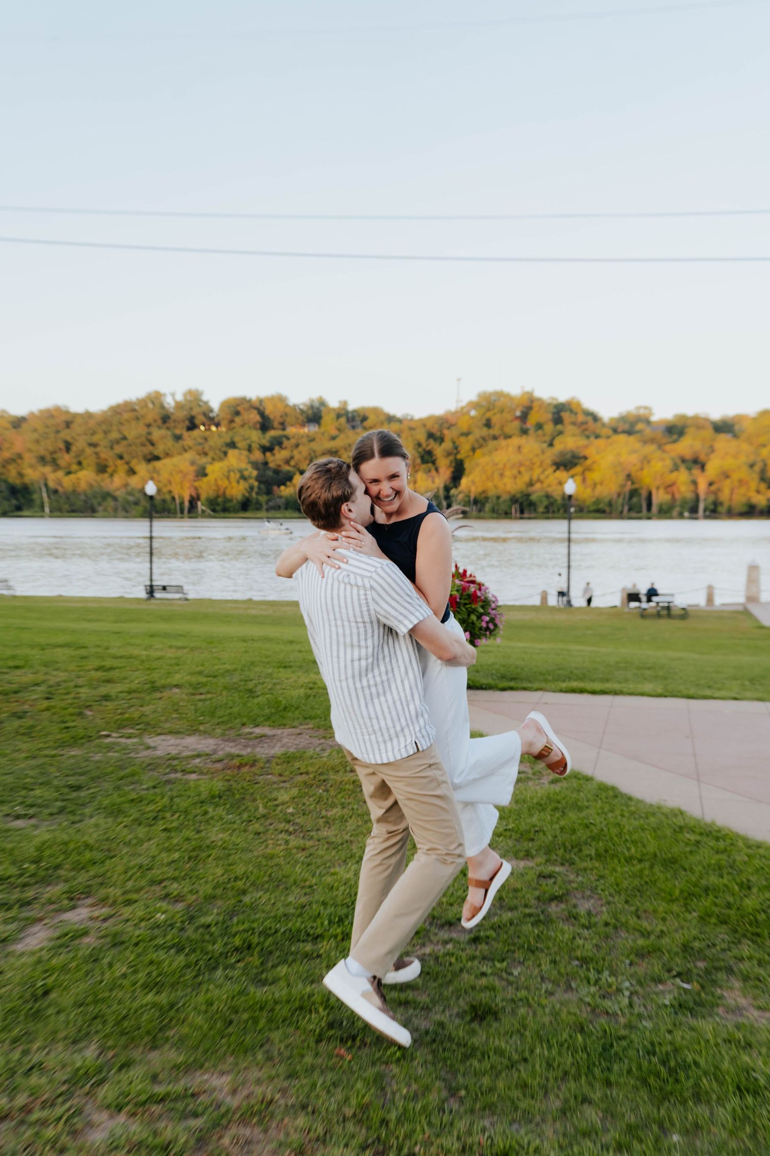 A couple dances playfully on a grassy lawn near a lake during golden hour, with one person being dipped dramatically.