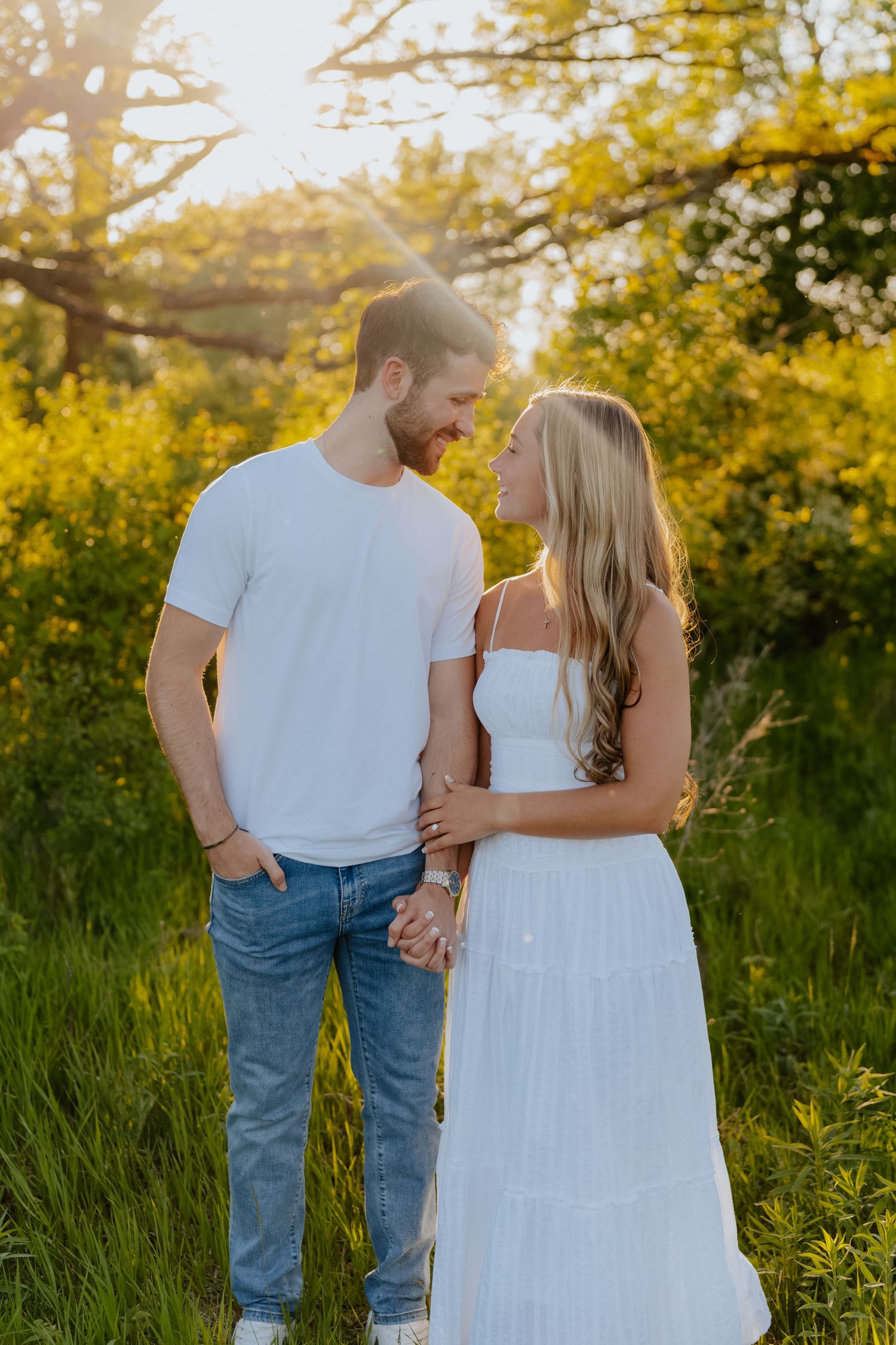 Couple in white outfits holding hands in a sunlit meadow with golden hour light filtering through green trees.