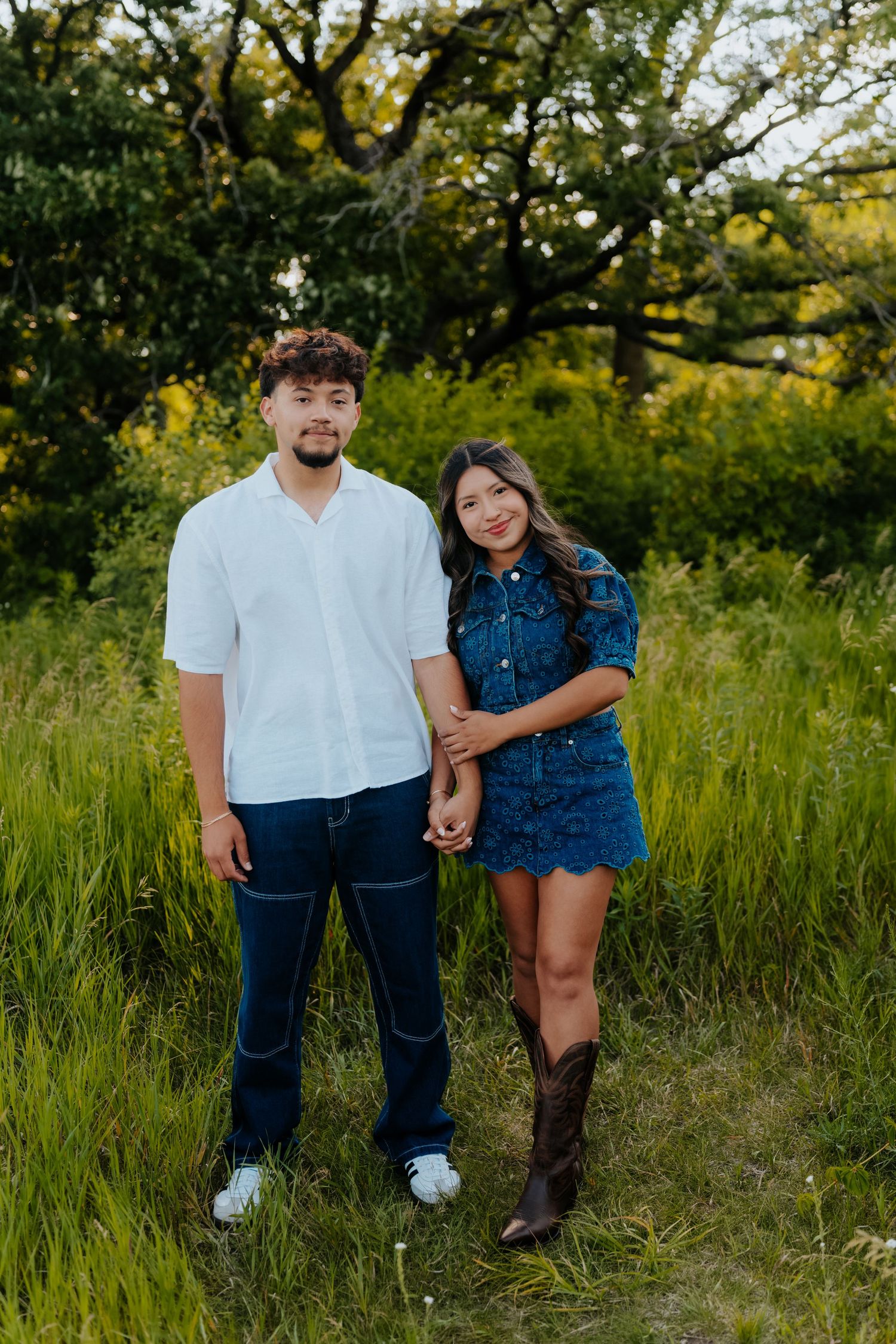 A young couple holds hands in a lush green field, man in white shirt and jeans, woman in blue denim dress and cowboy boots.
