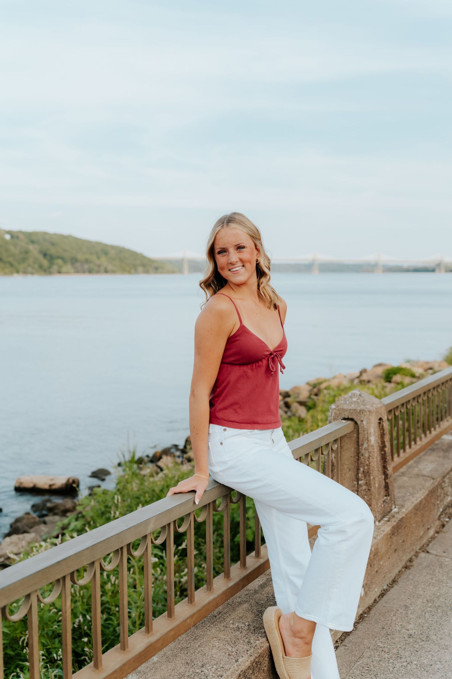 Young woman in red cami and white jeans smiling by a scenic waterfront railing on a sunny day.