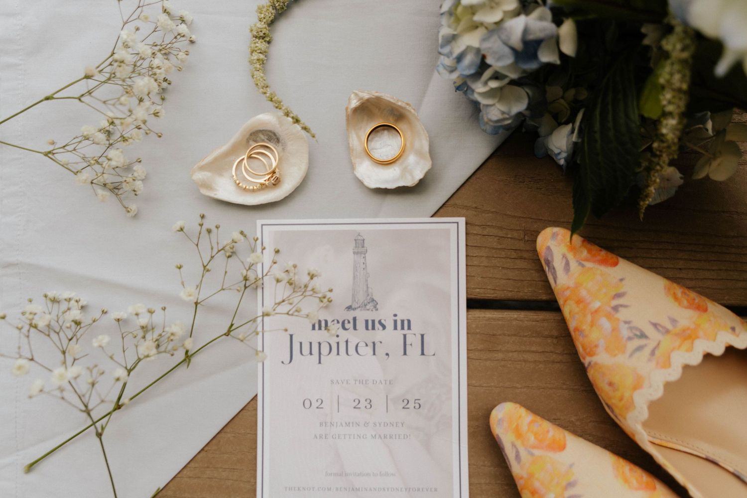 Wedding flat lay featuring invitation for Jupiter FL, gold rings, baby's breath, blue hydrangeas, and floral heels on wood.