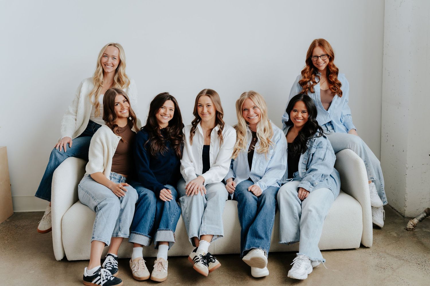 Seven women in casual denim outfits smiling together on a white couch in a bright studio setting.
