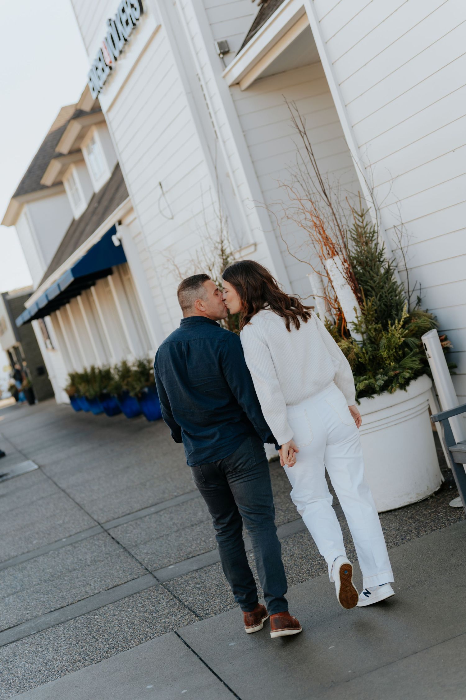Couple sharing a kiss while holding hands on a sidewalk outside a white building.