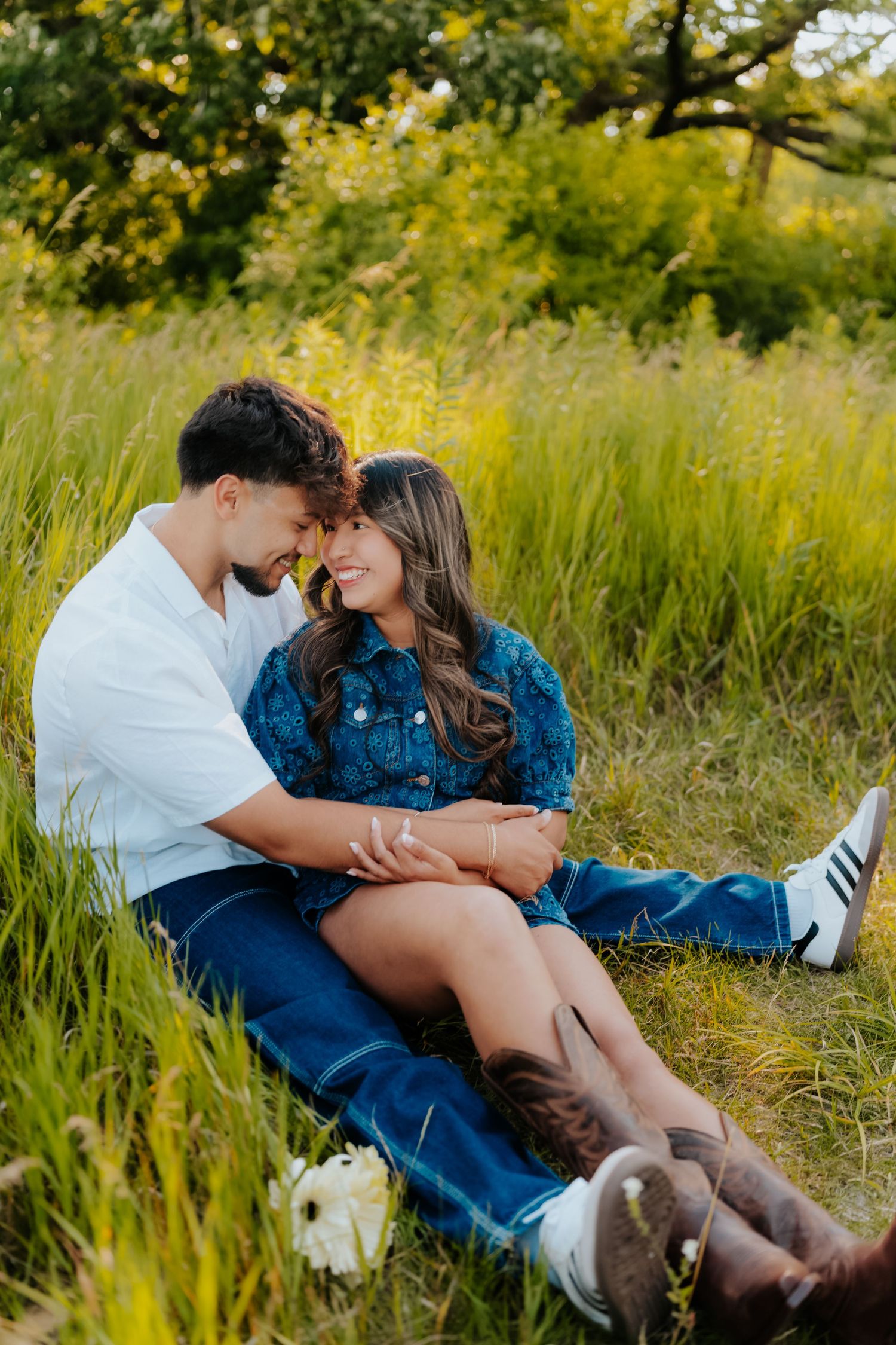 Couple sitting in green field, foreheads touching, smiling, wearing denim outfits, with wildflowers nearby.