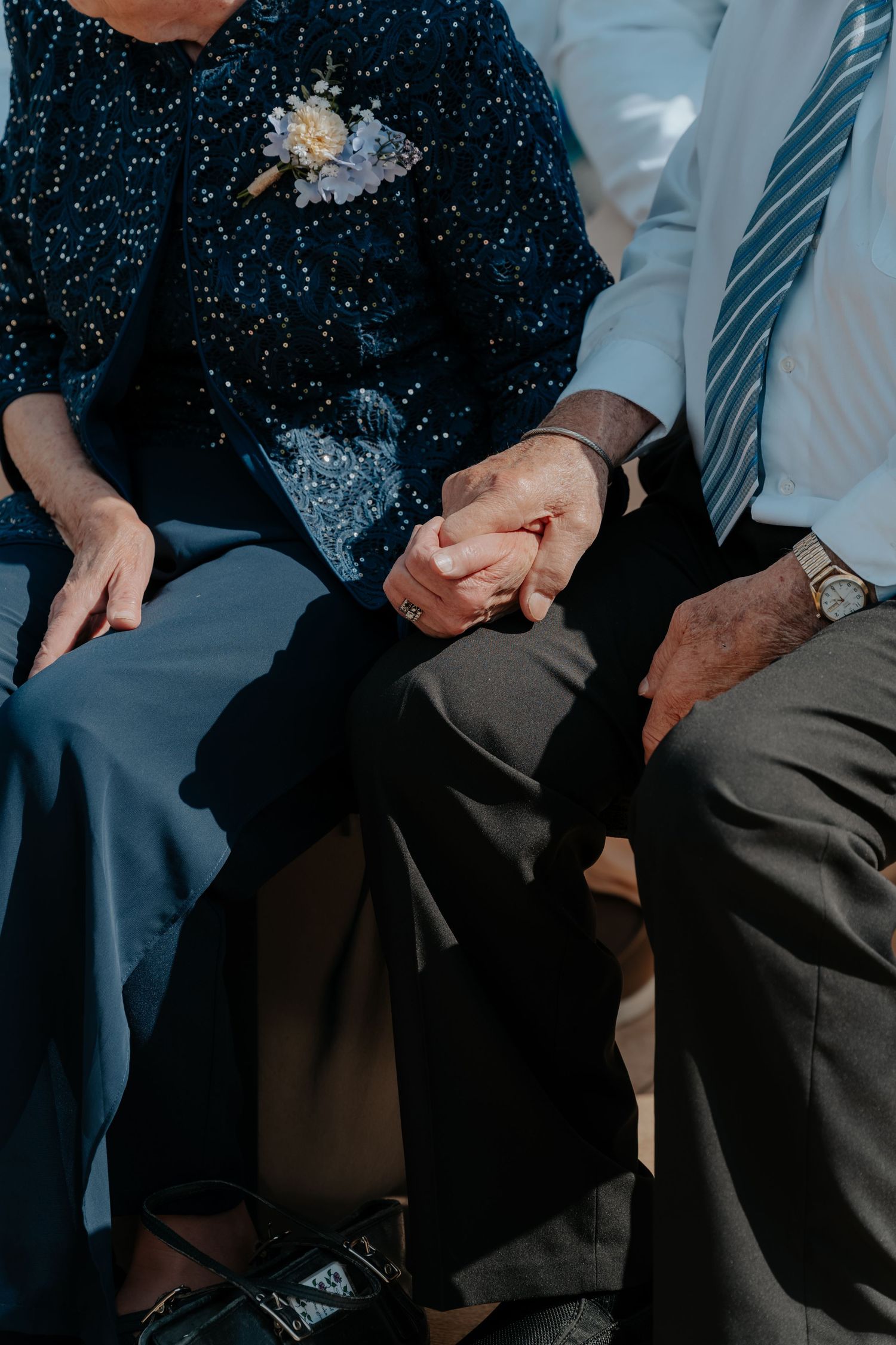 Elderly couple holding hands tenderly at wedding, woman wearing floral corsage, man in blue striped tie.