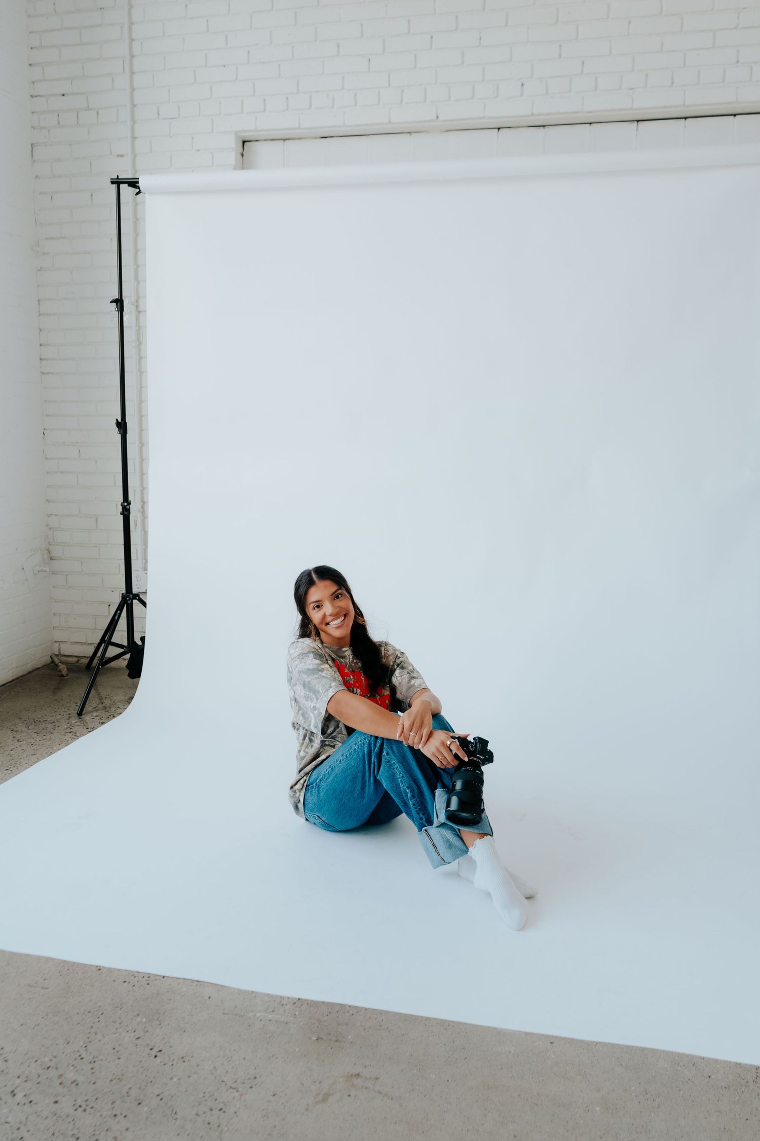 Smiling woman sitting on white backdrop in photography studio holding camera, wearing jeans and casual shirt.