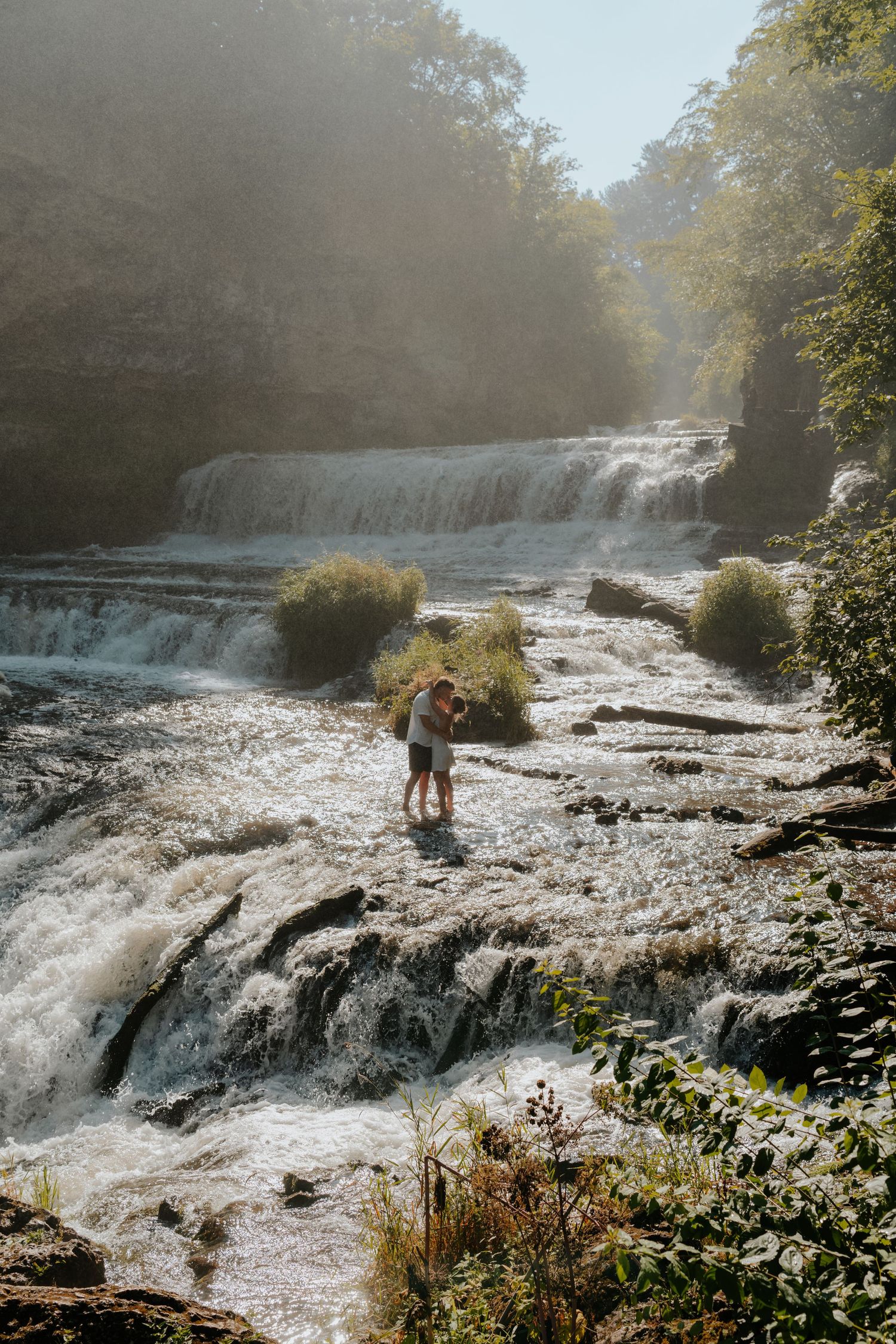 Couple embracing on rocky waterfall steps surrounded by lush green trees in soft morning light.