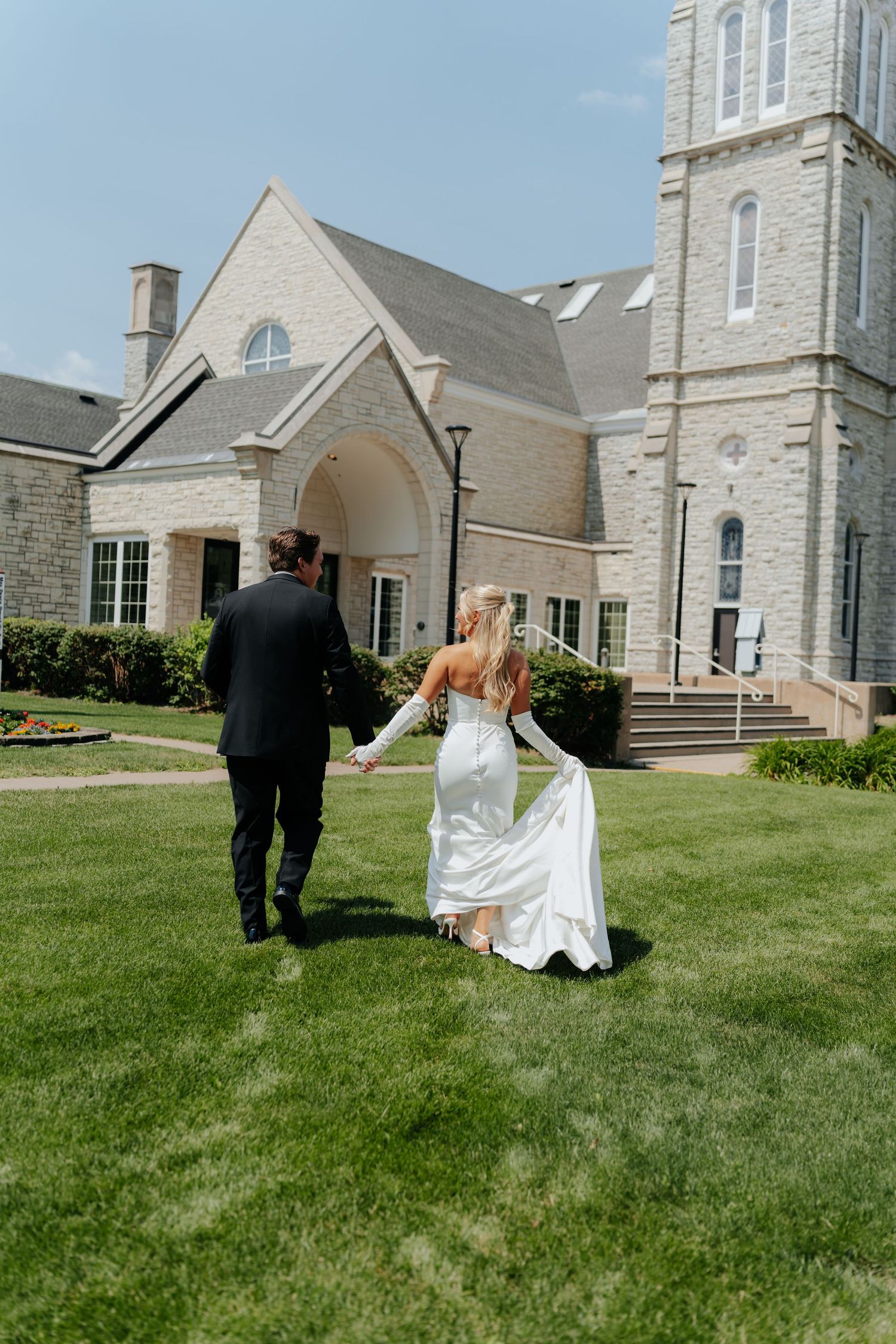 Bride and groom walking hand in hand across green lawn toward elegant stone church on sunny wedding day.