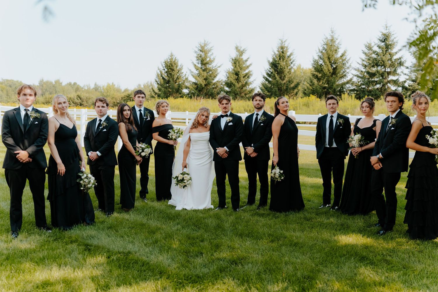 Wedding party in black attire poses on green lawn with evergreen trees, bride in white gown at center.