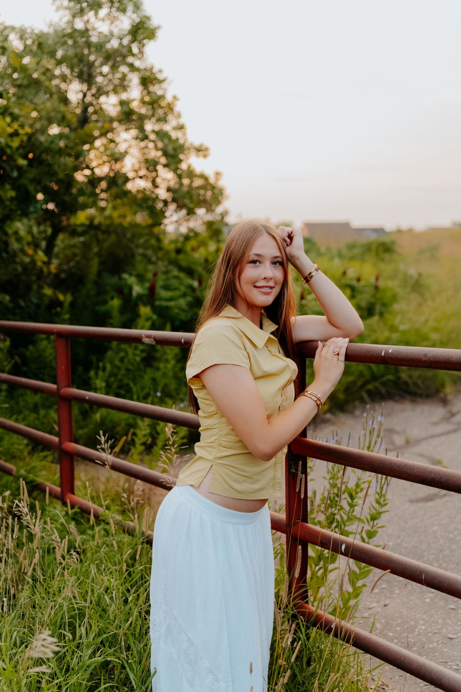 Young woman in yellow top and light blue skirt poses by rustic metal fence with lush green trees at golden hour.