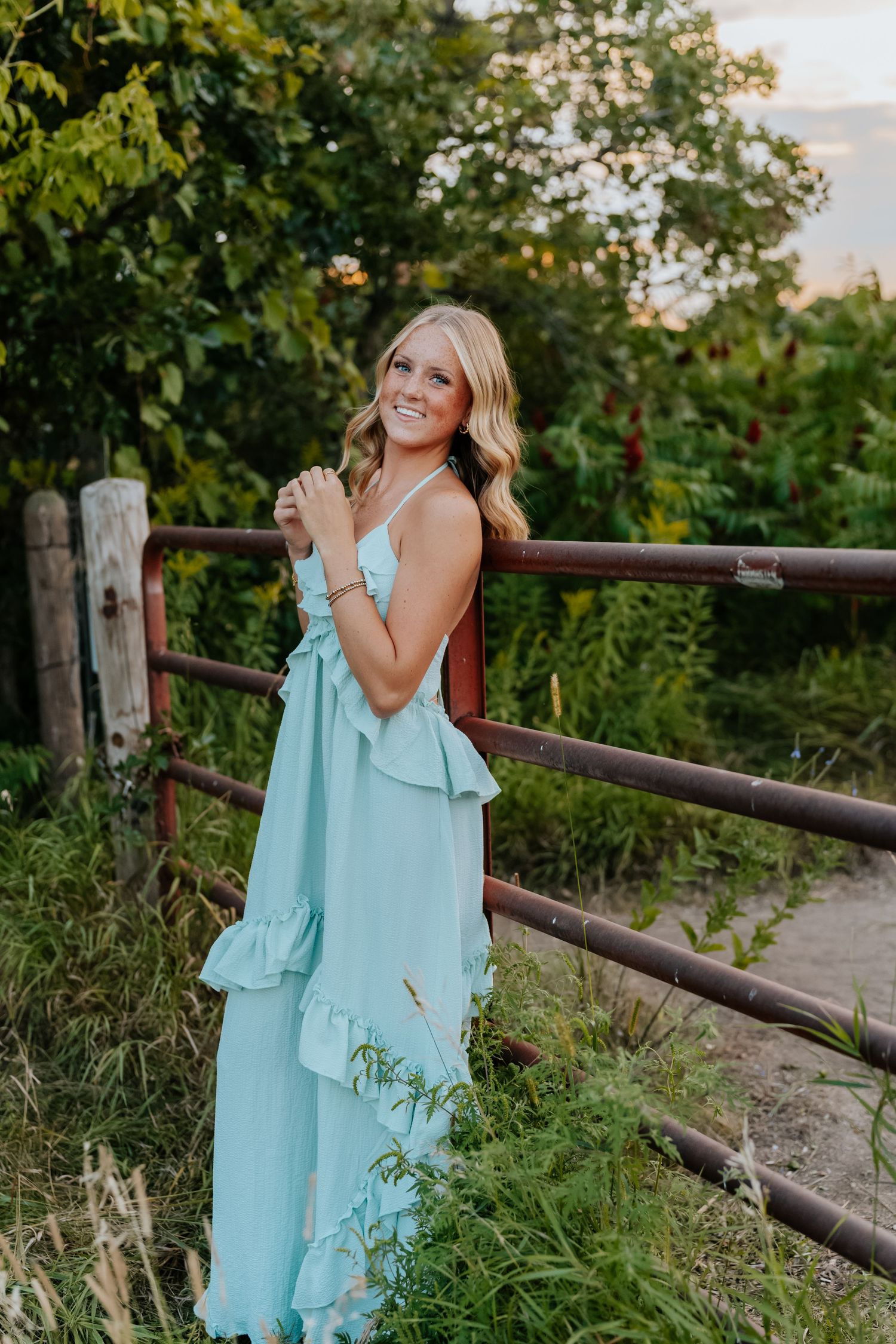 Young woman in mint ruffle maxi dress smiling by rustic wooden fence surrounded by lush greenery at golden hour.