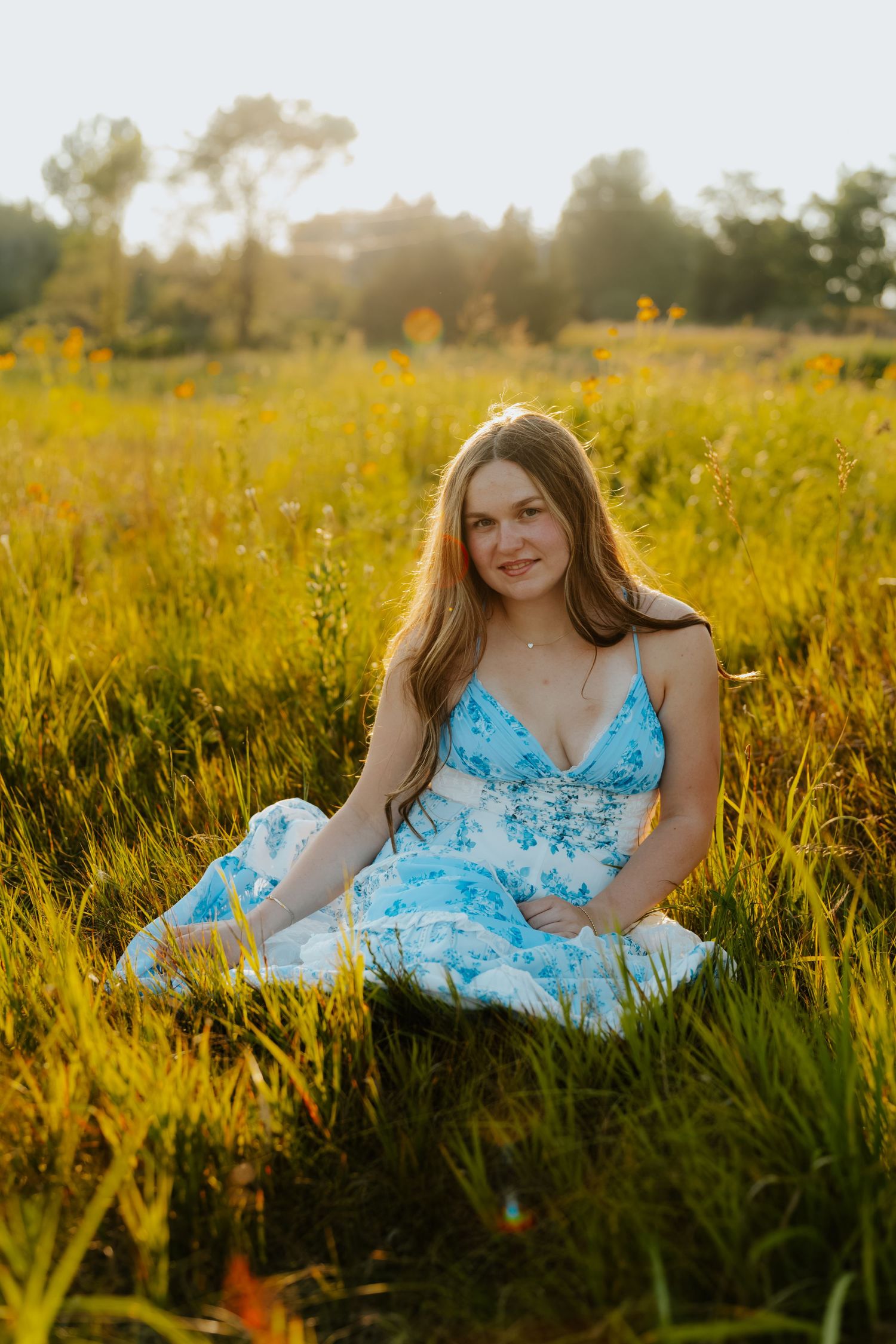 Young woman in blue floral dress sitting in sunny wildflower meadow, golden hour light, outdoor portrait.