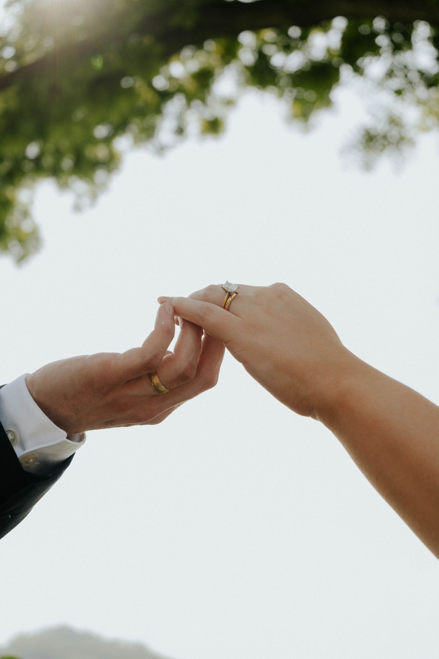 Close-up of hands with wedding rings reaching towards each other against a bright sky.