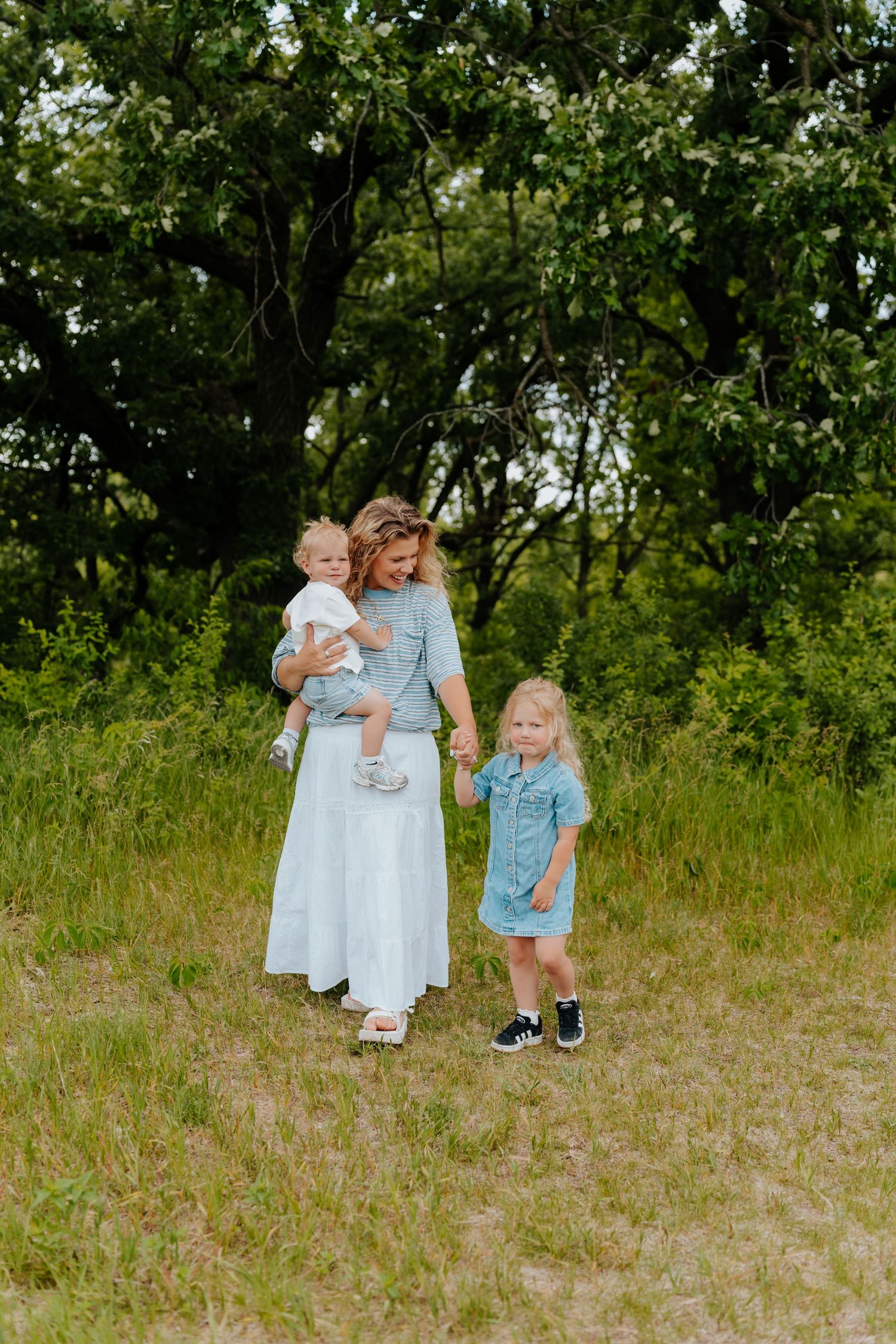 A mother in a light blue dress holds her baby while walking hand-in-hand with her young daughter outdoors.