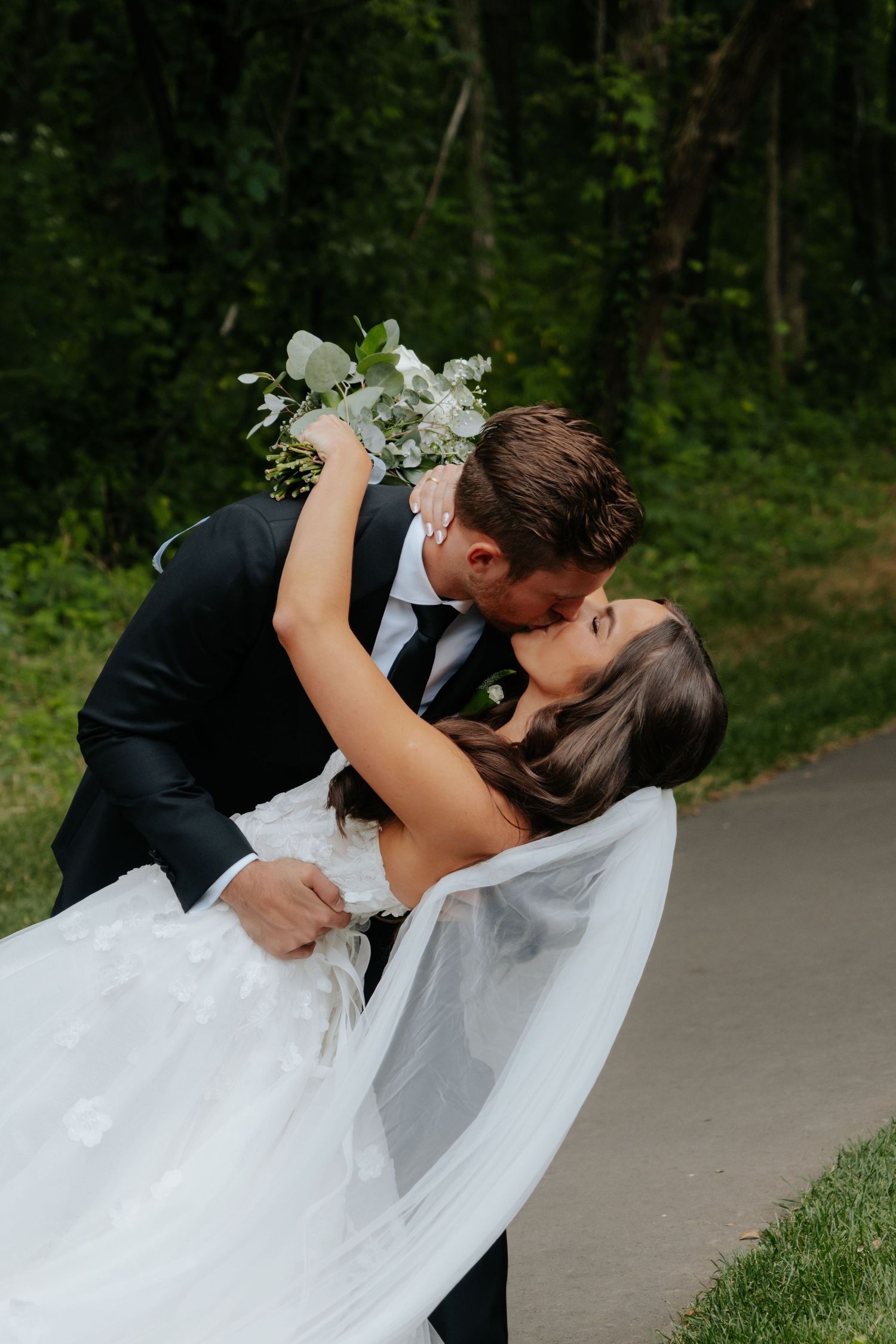 Groom in black suit dips bride in white gown for a romantic kiss, holding a white floral bouquet outdoors.