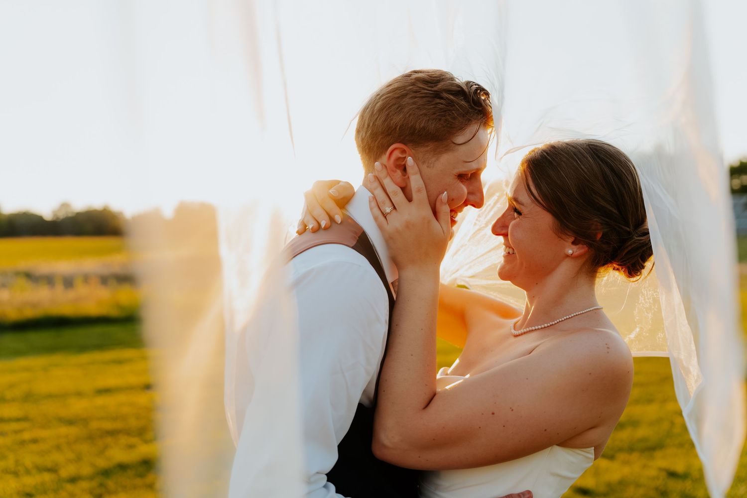 A bride and groom share a romantic moment under a flowing veil at golden hour in a sunlit outdoor field.