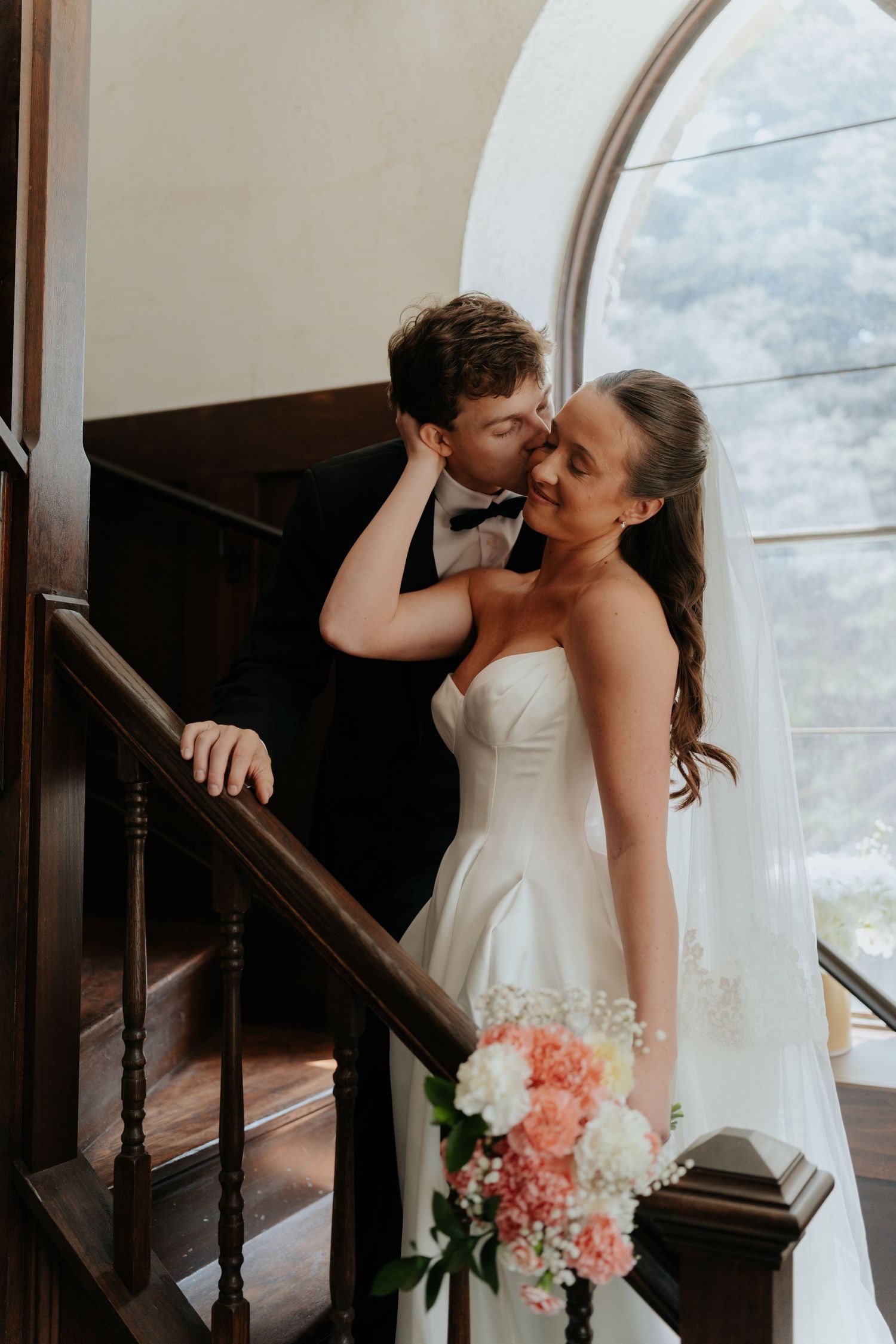Bride and groom share a tender kiss on elegant wooden staircase, bride holding pink and white bouquet wearing strapless gown.