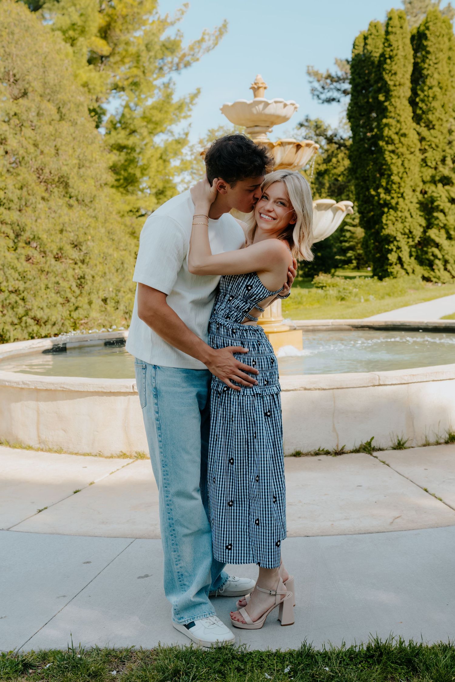 Couple embracing and kissing by a stone fountain in a lush garden, woman in blue gingham dress smiling.