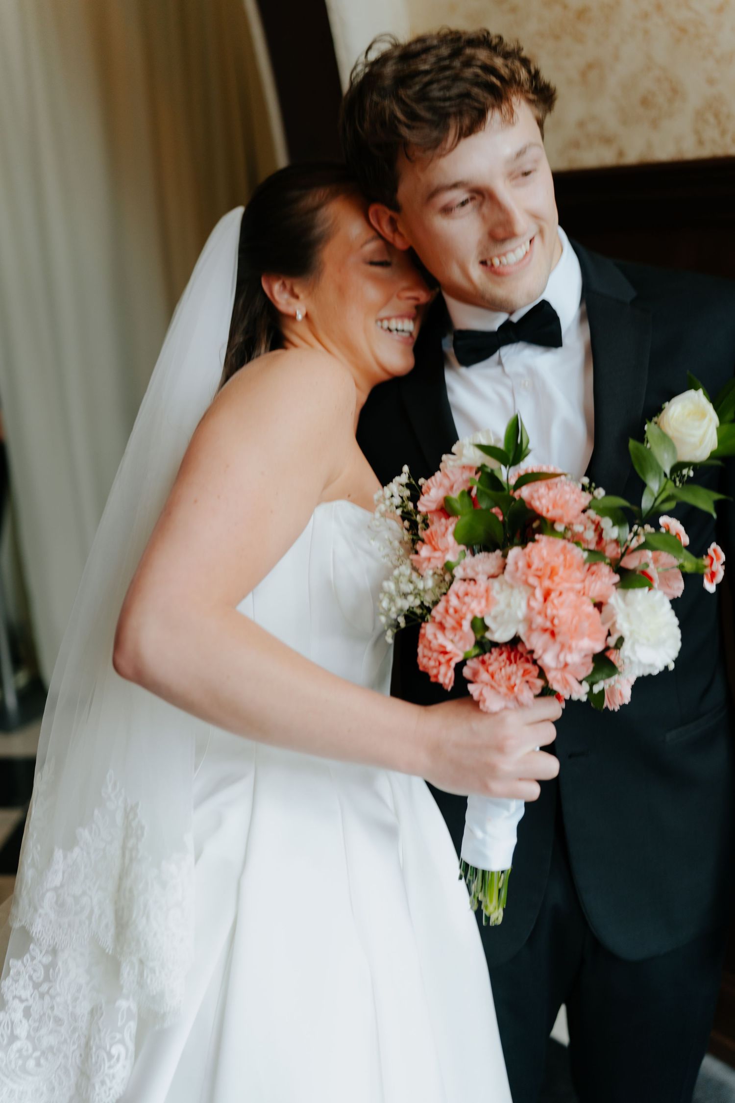 Smiling bride and groom holding a pink and white bouquet, posing together in elegant wedding attire.