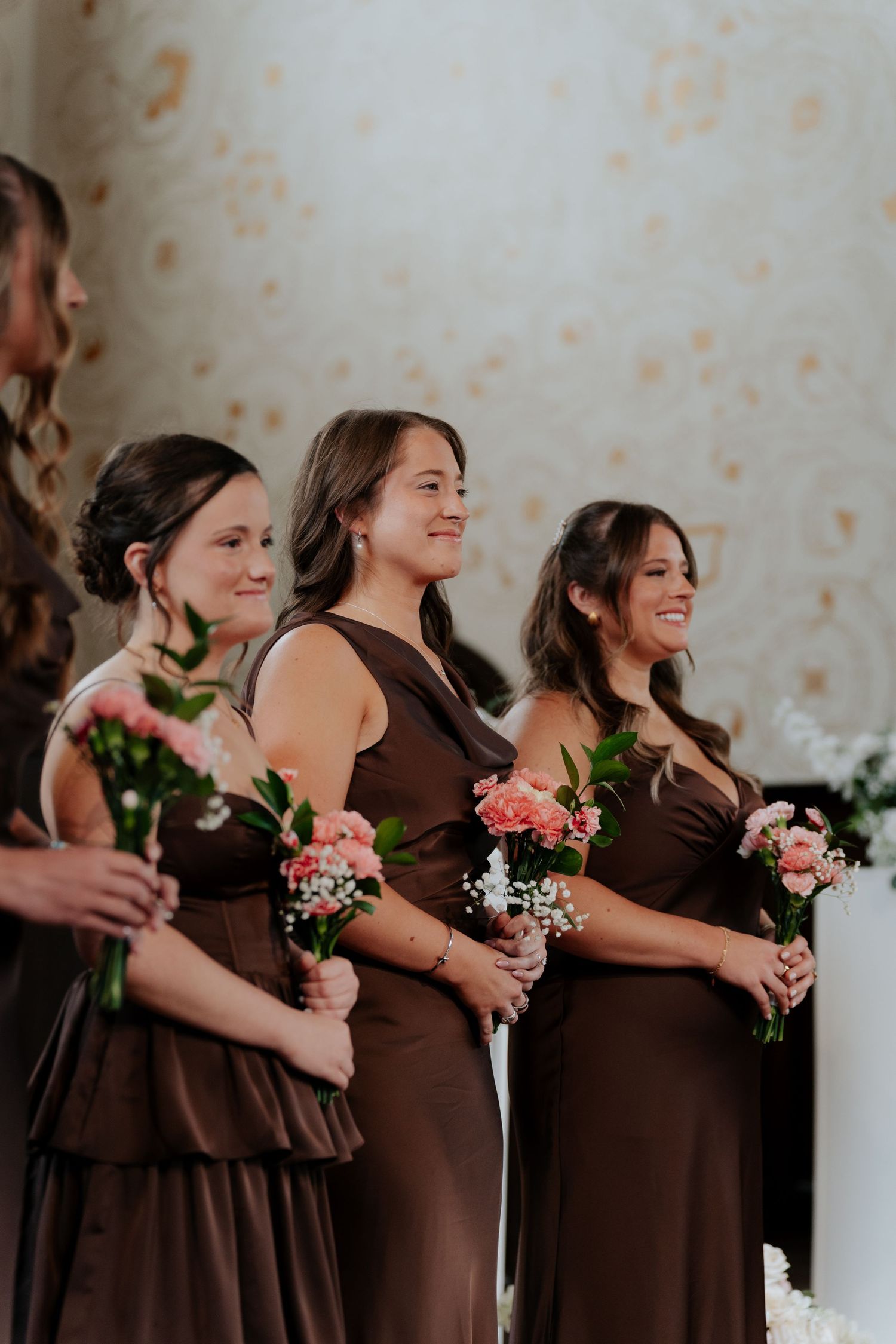 Bridesmaids in brown dresses holding pink floral bouquets stand together smiling during a wedding ceremony.