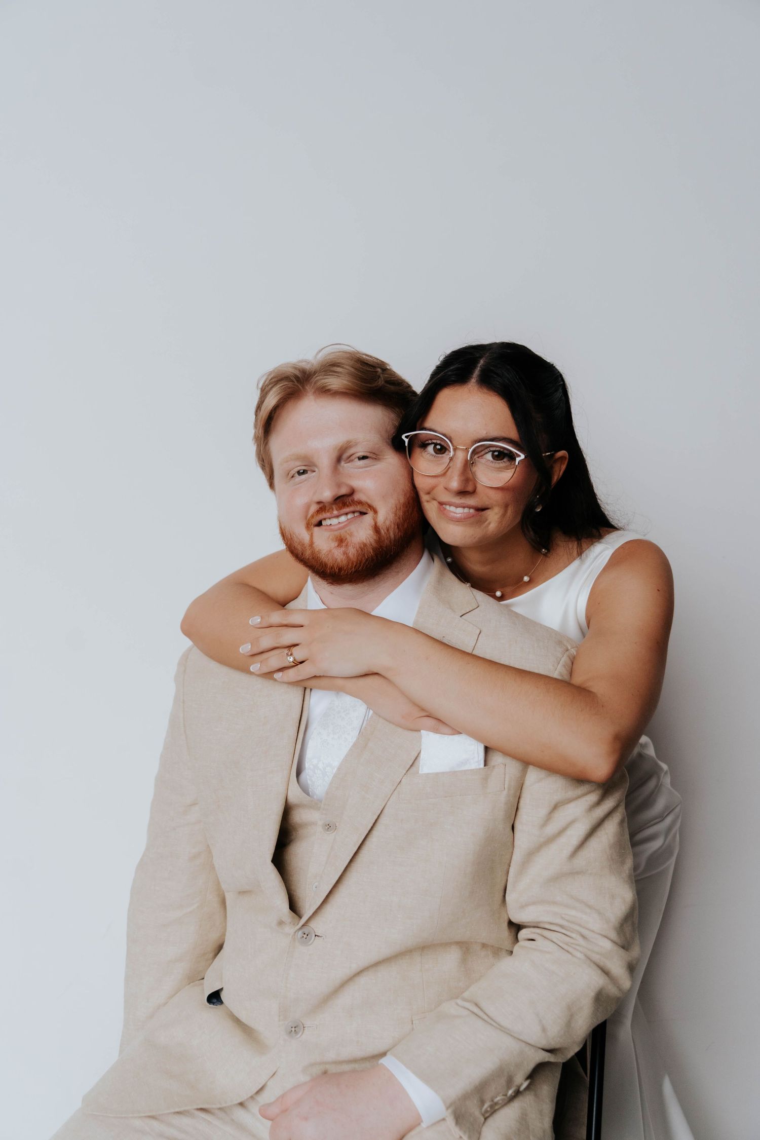A smiling couple poses together, woman with glasses hugging a red-bearded man in a beige suit against a white background.