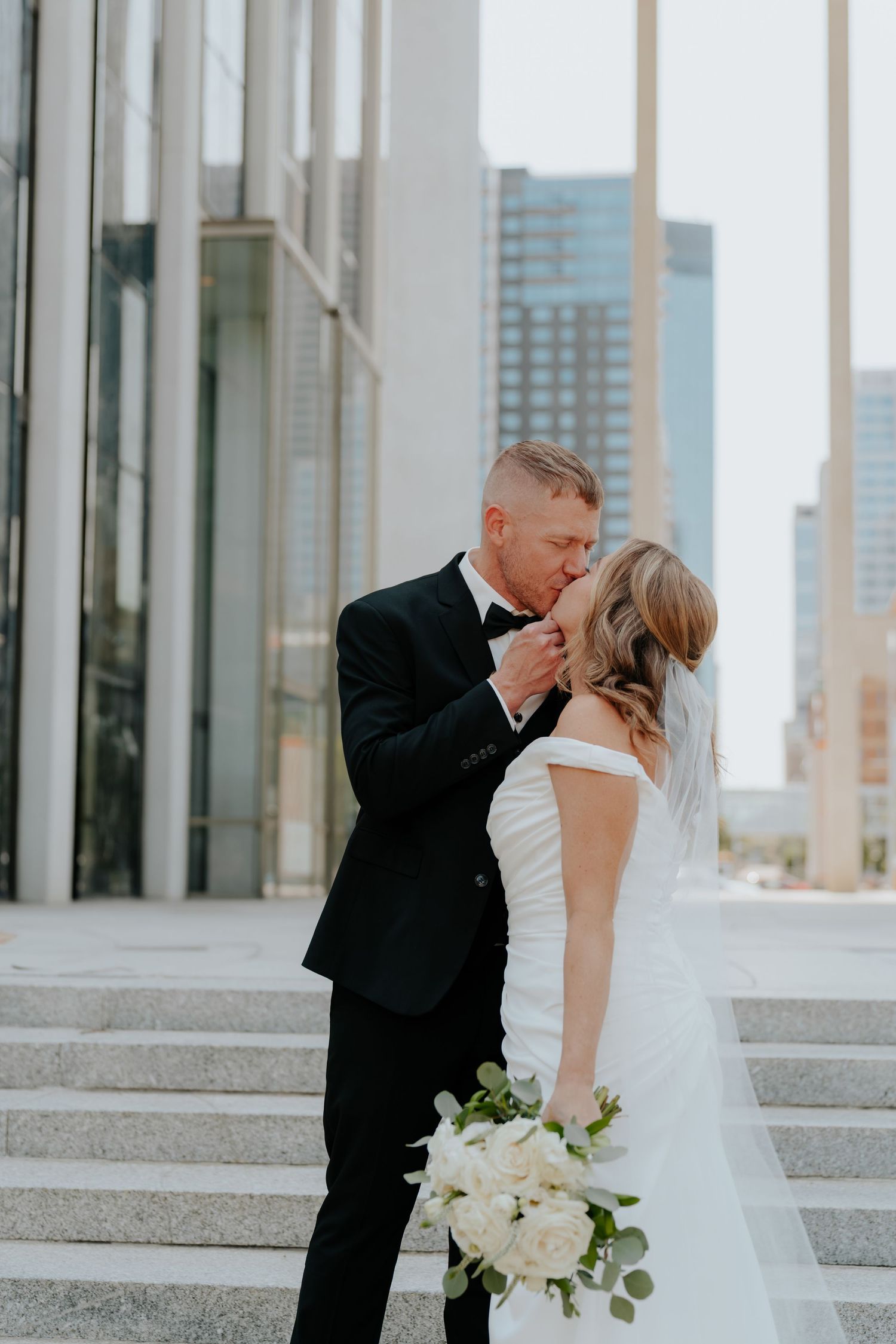 Bride and groom share a kiss on city steps, she holds white bouquet, he wears black tuxedo, urban skyline backdrop.
