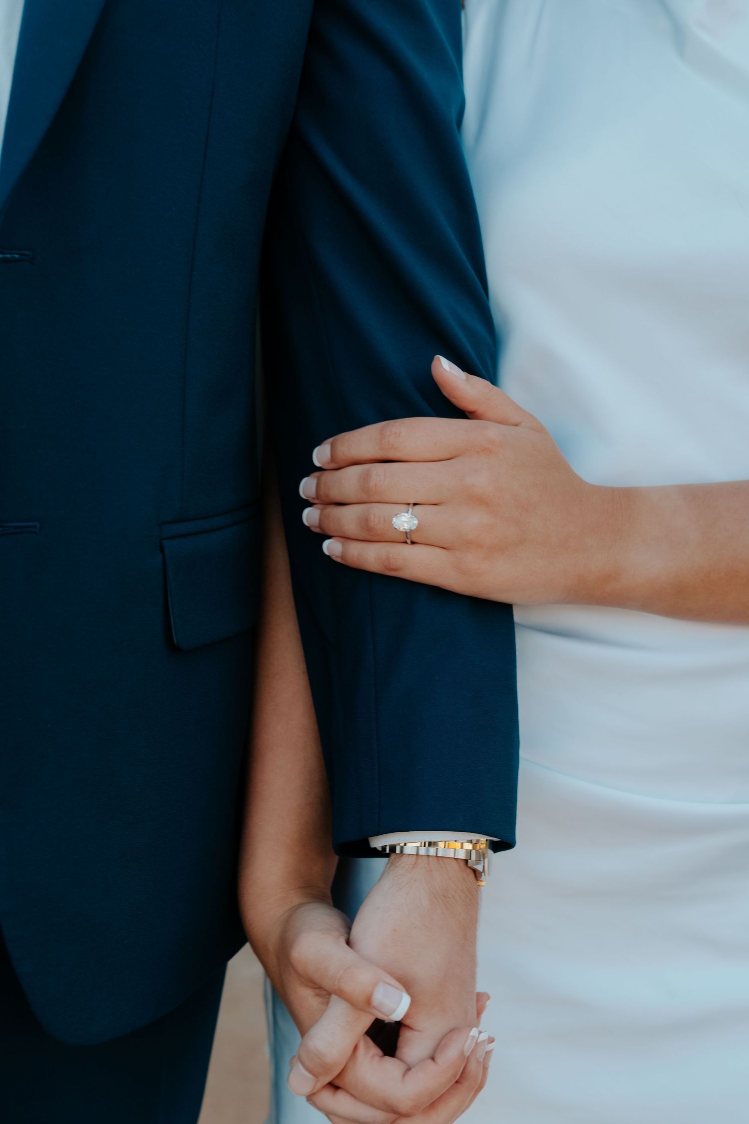 Bride in white dress holds groom's hand, showcasing elegant oval engagement ring and gold bracelet.
